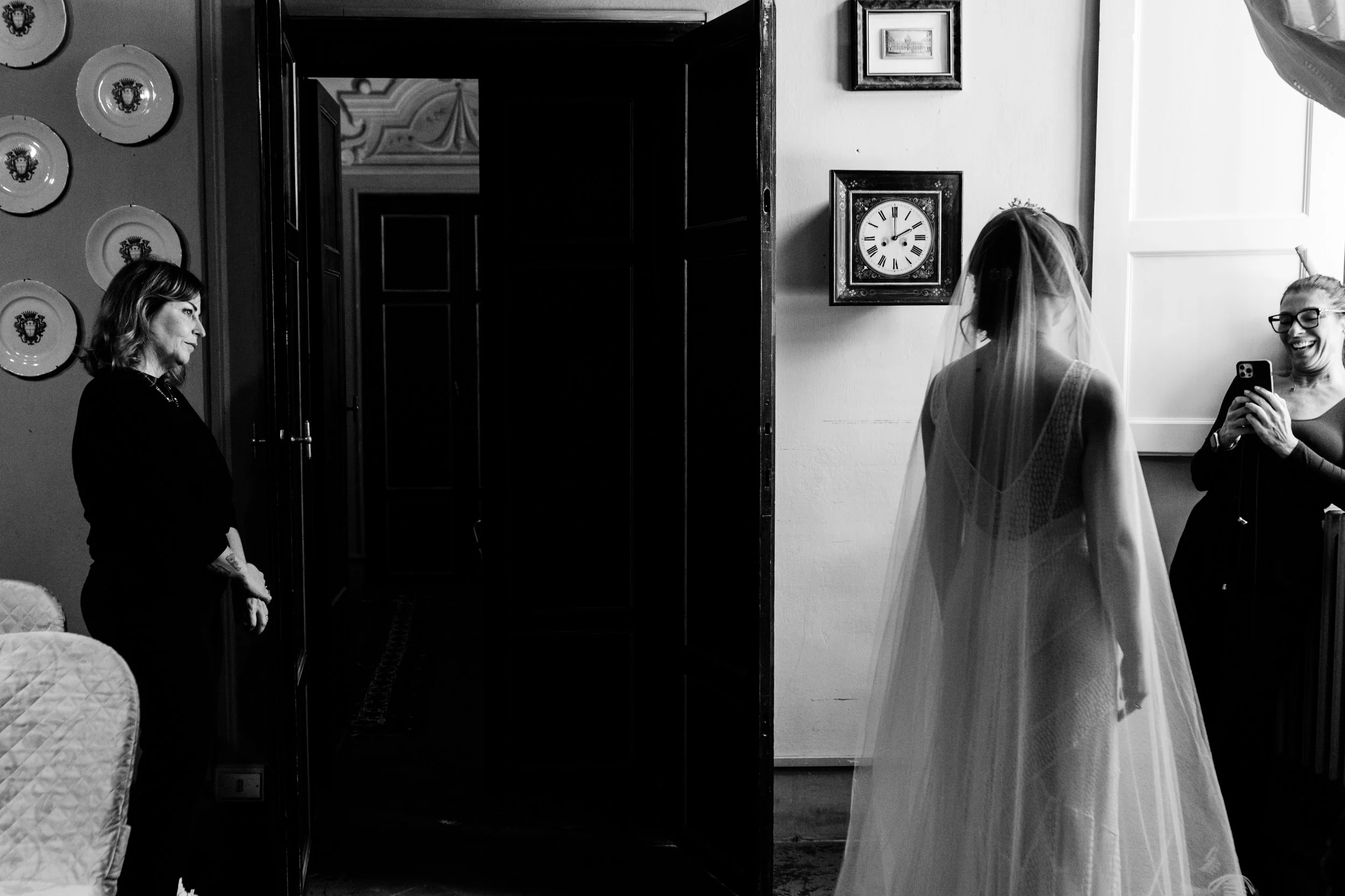 Una donna con abito da sposa e velo si trova davanti a due donne, una che sorride e le fa foto, in una stanza con pareti bianche e orologio. Altri dettagli includono piatti decorativi e dipinti alle pareti. Villa di Corliano