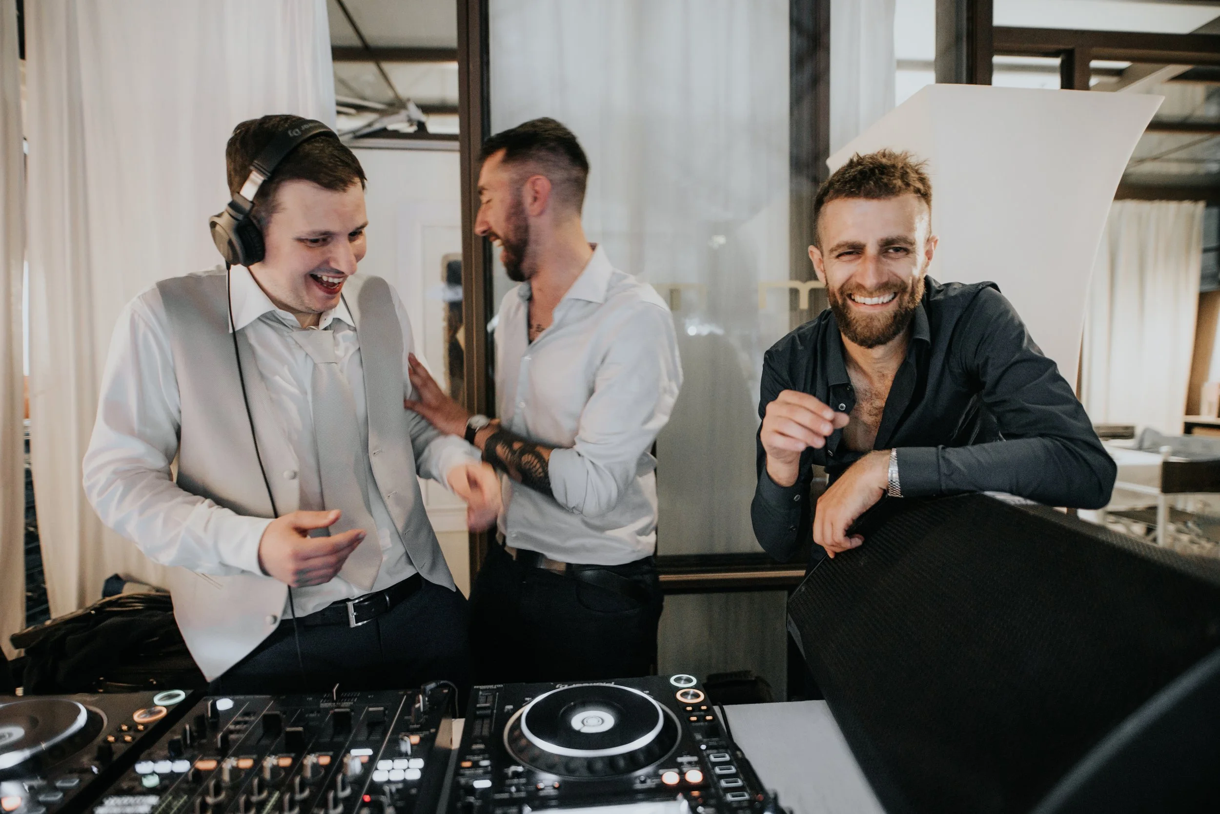 Tre uomini sorridenti in un locale, uno con cuffie e DJ con attrezzatura musicale, gli altri due che ridono e si abbracciano.