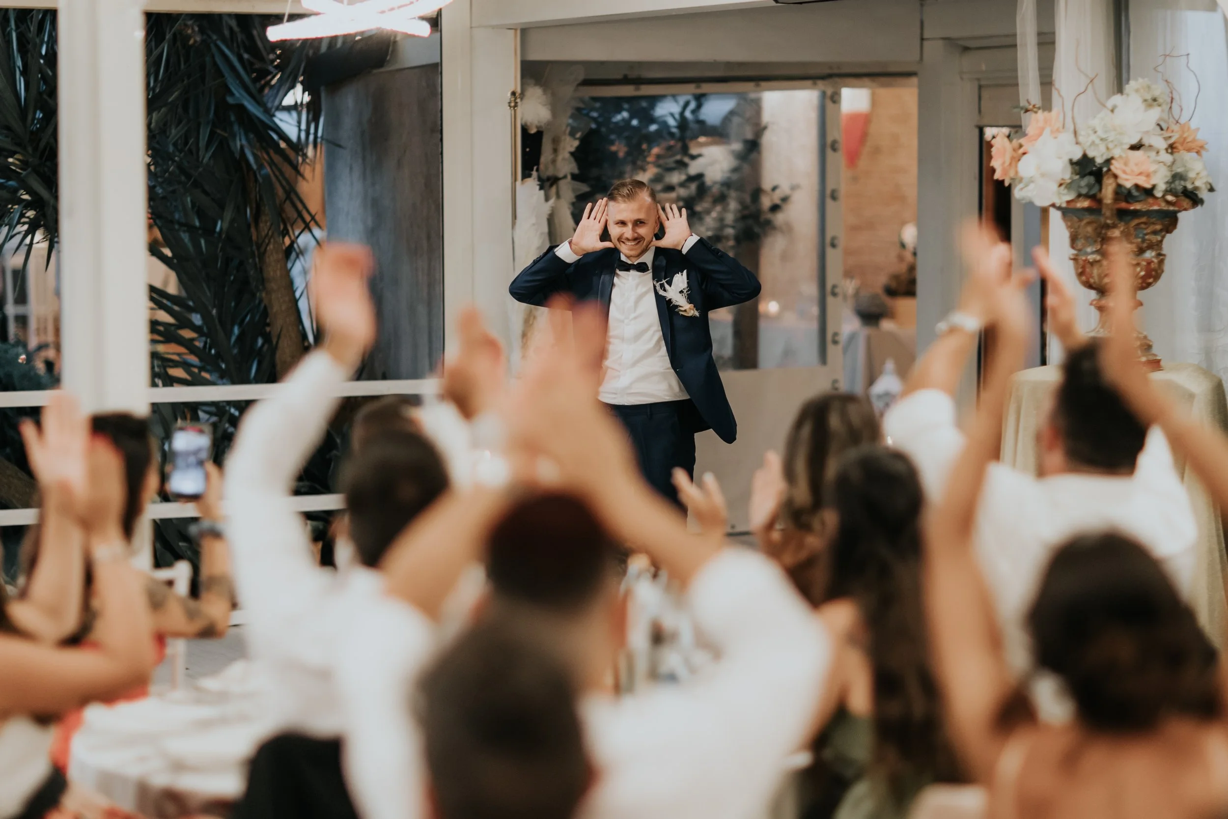 Lo sposo sorride mentre fa un discorso o una battuta davanti a ospiti seduti a un ricevimento di matrimonio, con le mani vicino alle orecchie, in un ambiente elegante decorato con fiori.