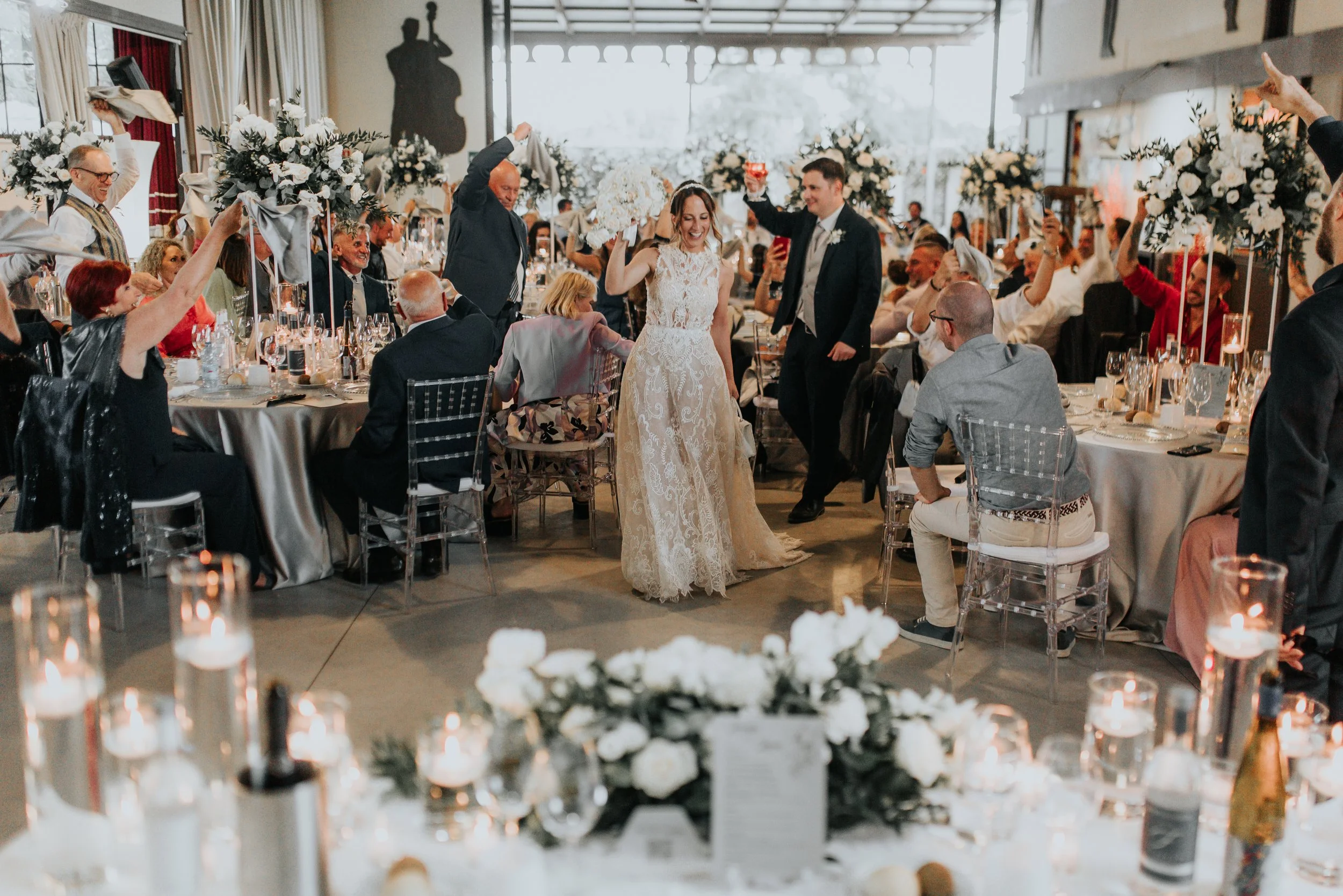 Una coppia di sposi balla felice al centro di una sala da pranzo elegante circondata da amici e parenti, con decorazioni floreali e candele.