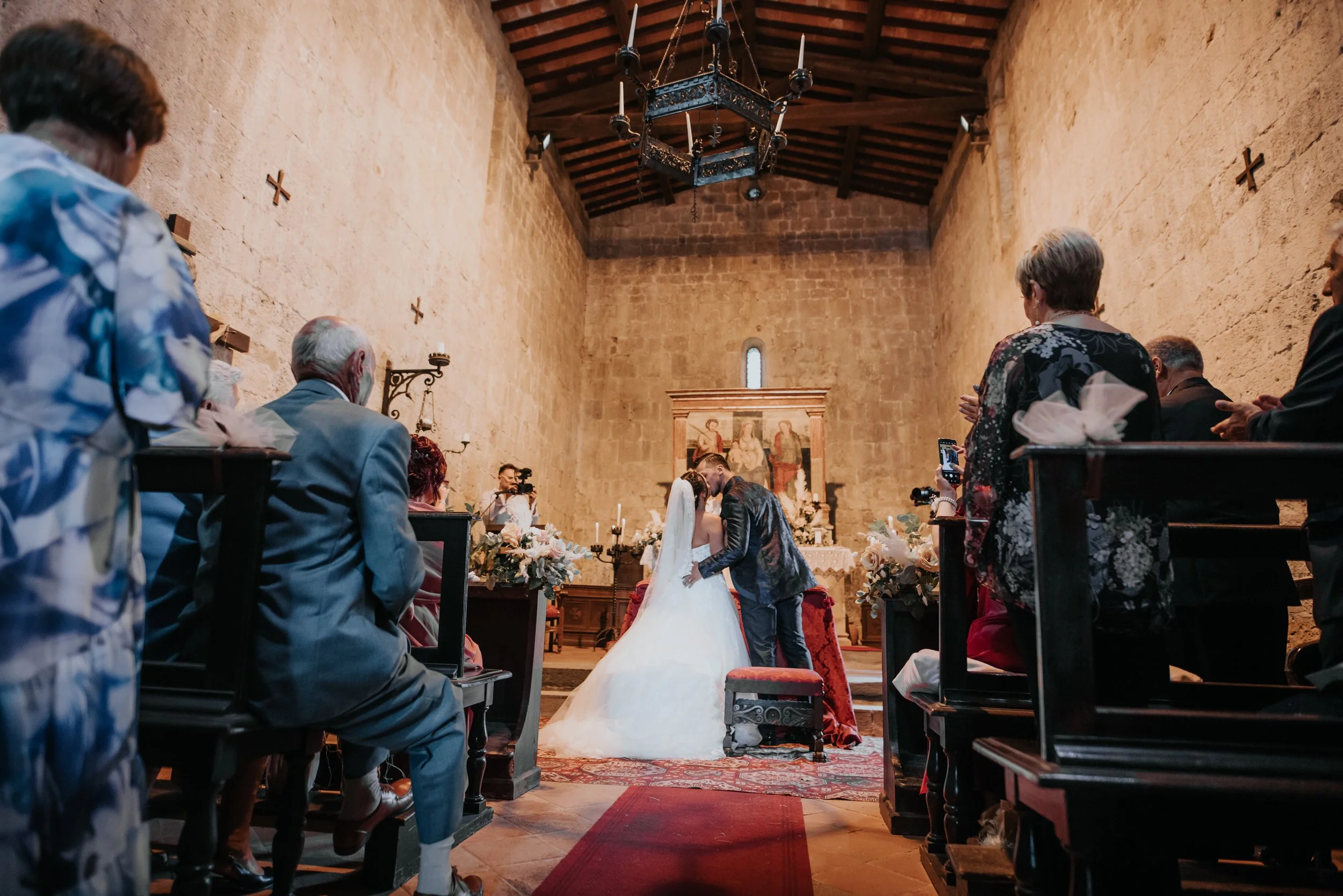 Una coppia di sposi si bacia davanti all'altare in una chiesa in pietra, circondati da amici e parenti seduti e che scattano foto.