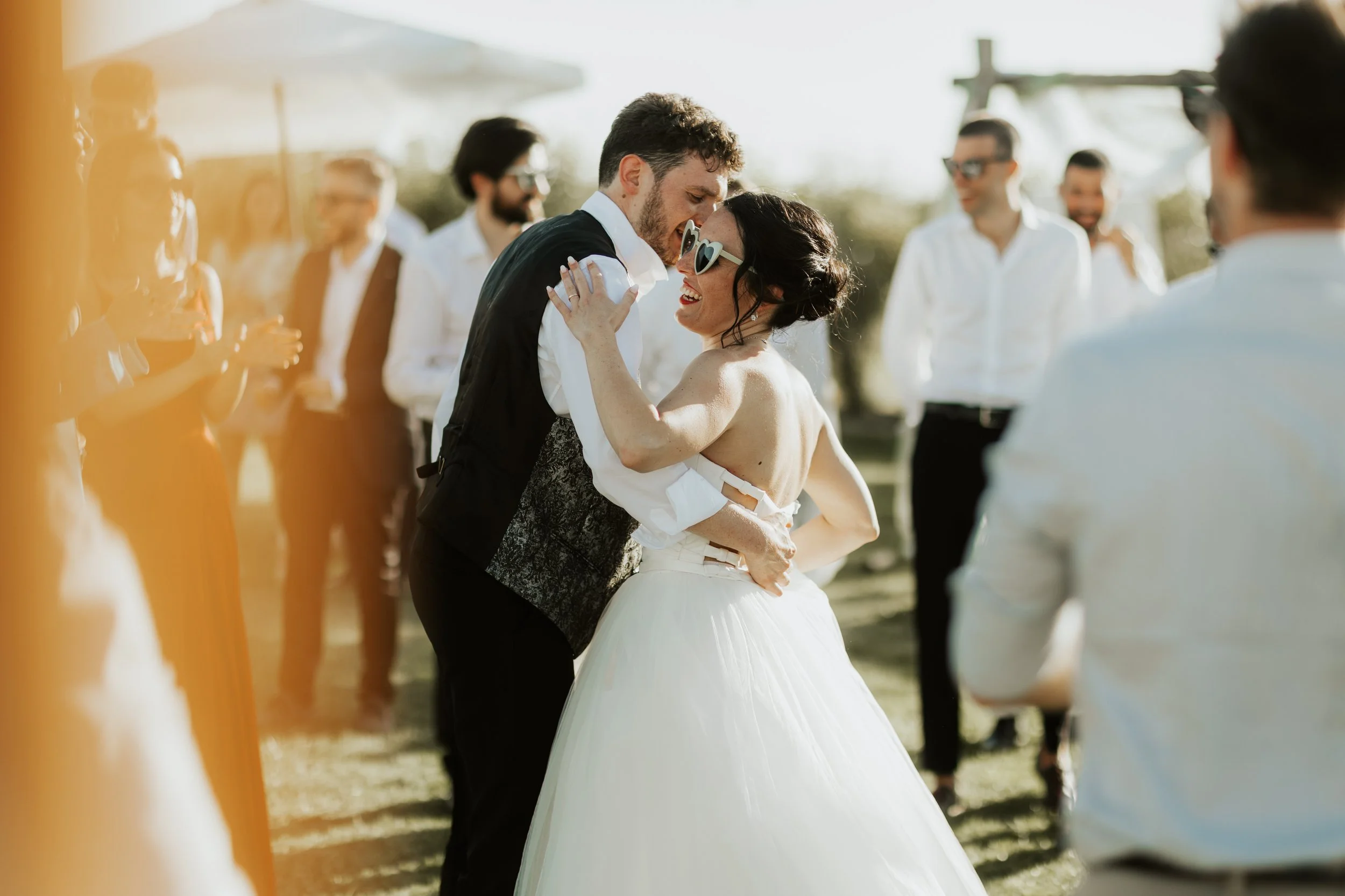 Una coppia di sposi che balla e ride durante il loro matrimonio all'aperto, circondati da amici e parenti.