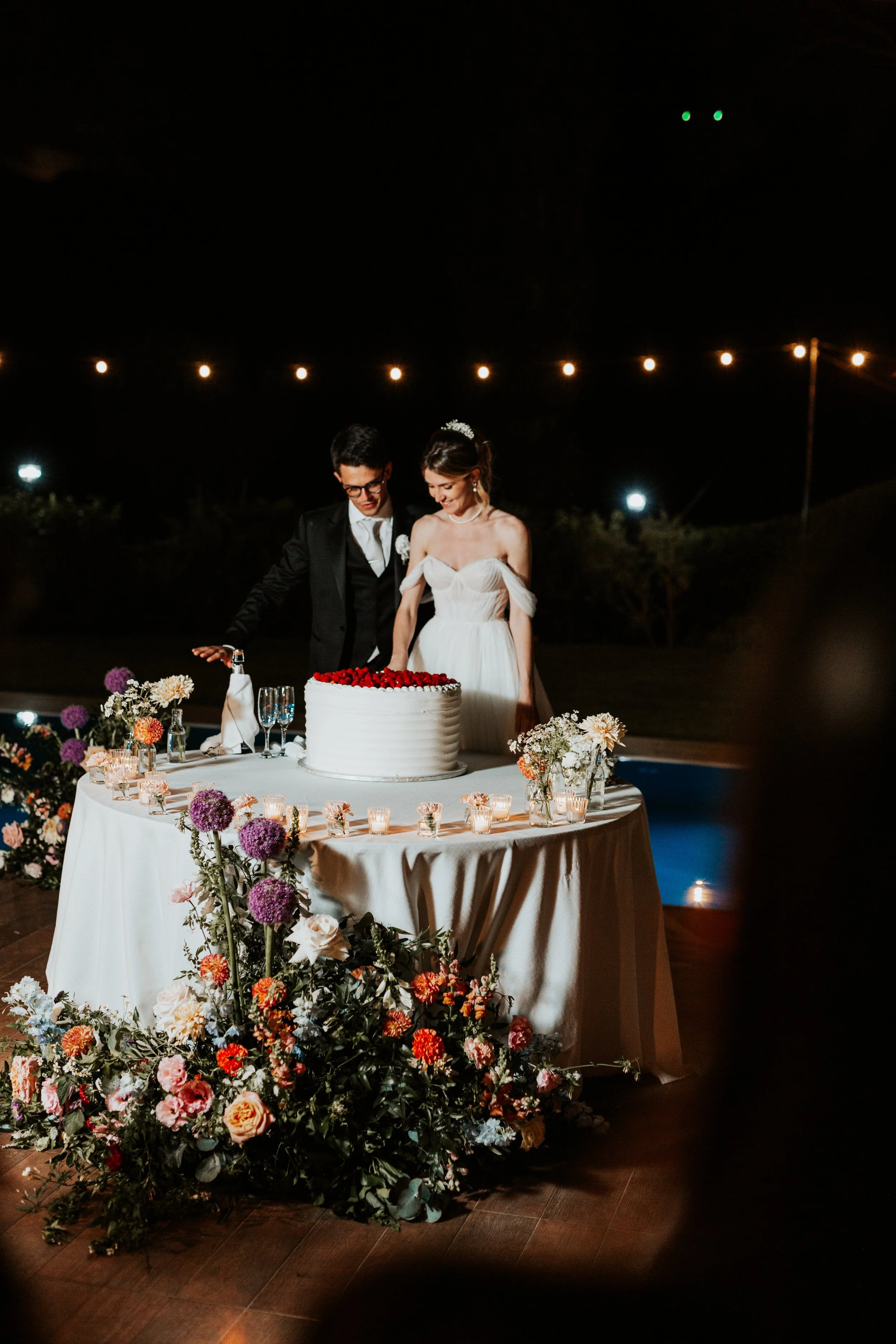 Una coppia di sposi si diverte davanti a una torta nuziale decorata con frutti di bosco, circondata da fiori colorati e candele, in un ambiente notturno con luci a festa.