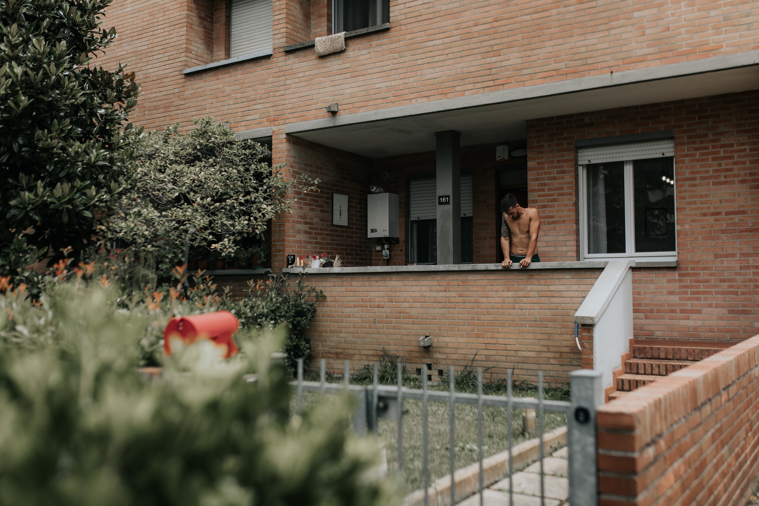 Uomo nudo con tatuaggio sulla braccia che si appoggia al parapetto di un balcone di un edificio in mattoni, con una finestra e una porta visibili sullo sfondo.