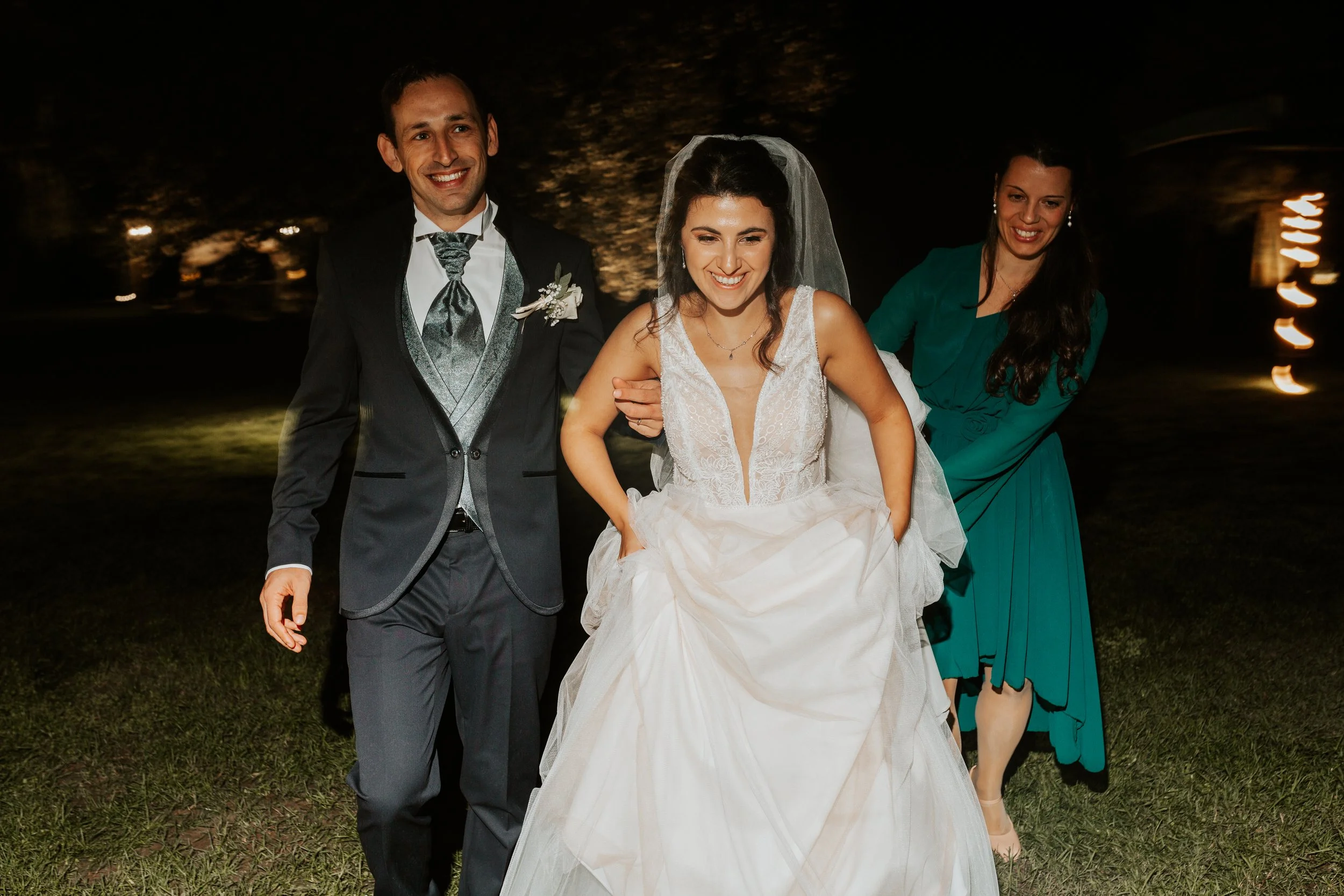 Una coppia di sposi e un'amica sorridenti camminano all'aperto di notte, con alberi e luci sfocate sullo sfondo.