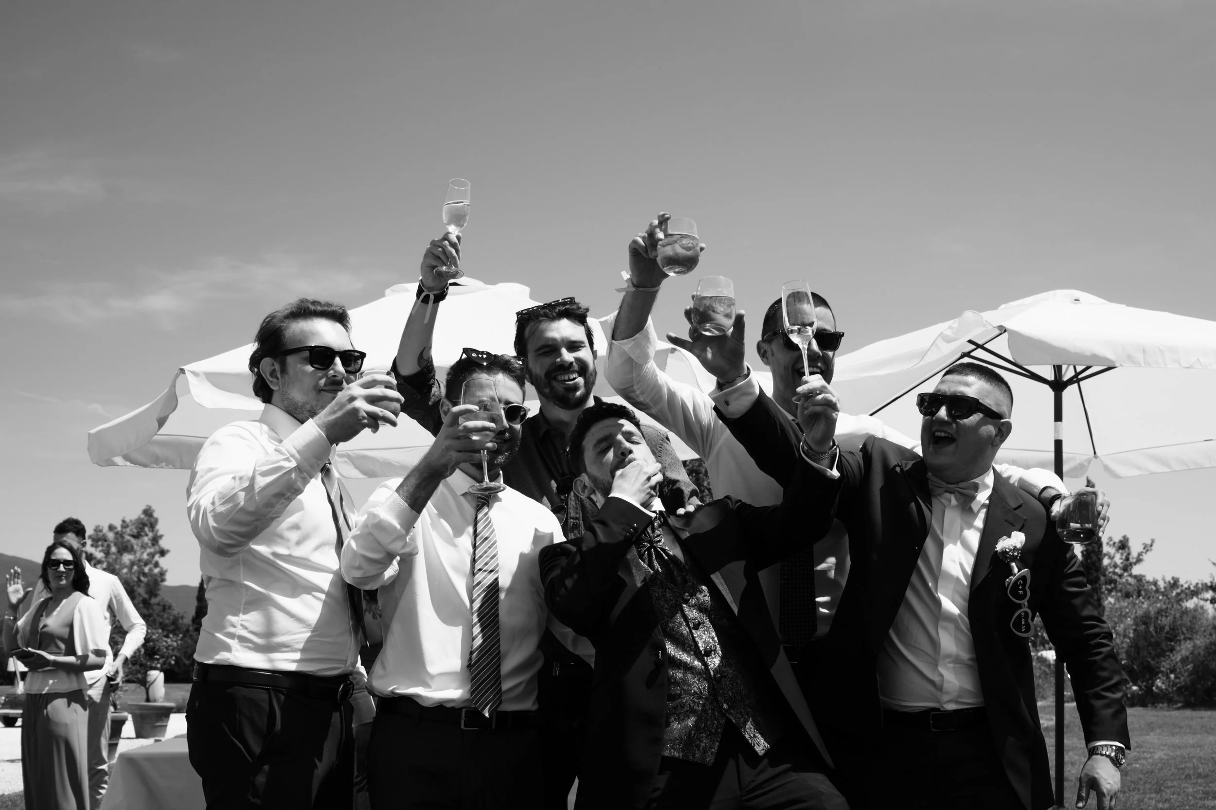 Gruppo di persone che brindano con bicchieri di vino all'aperto, sotto ombrelloni, durante una celebrazione.