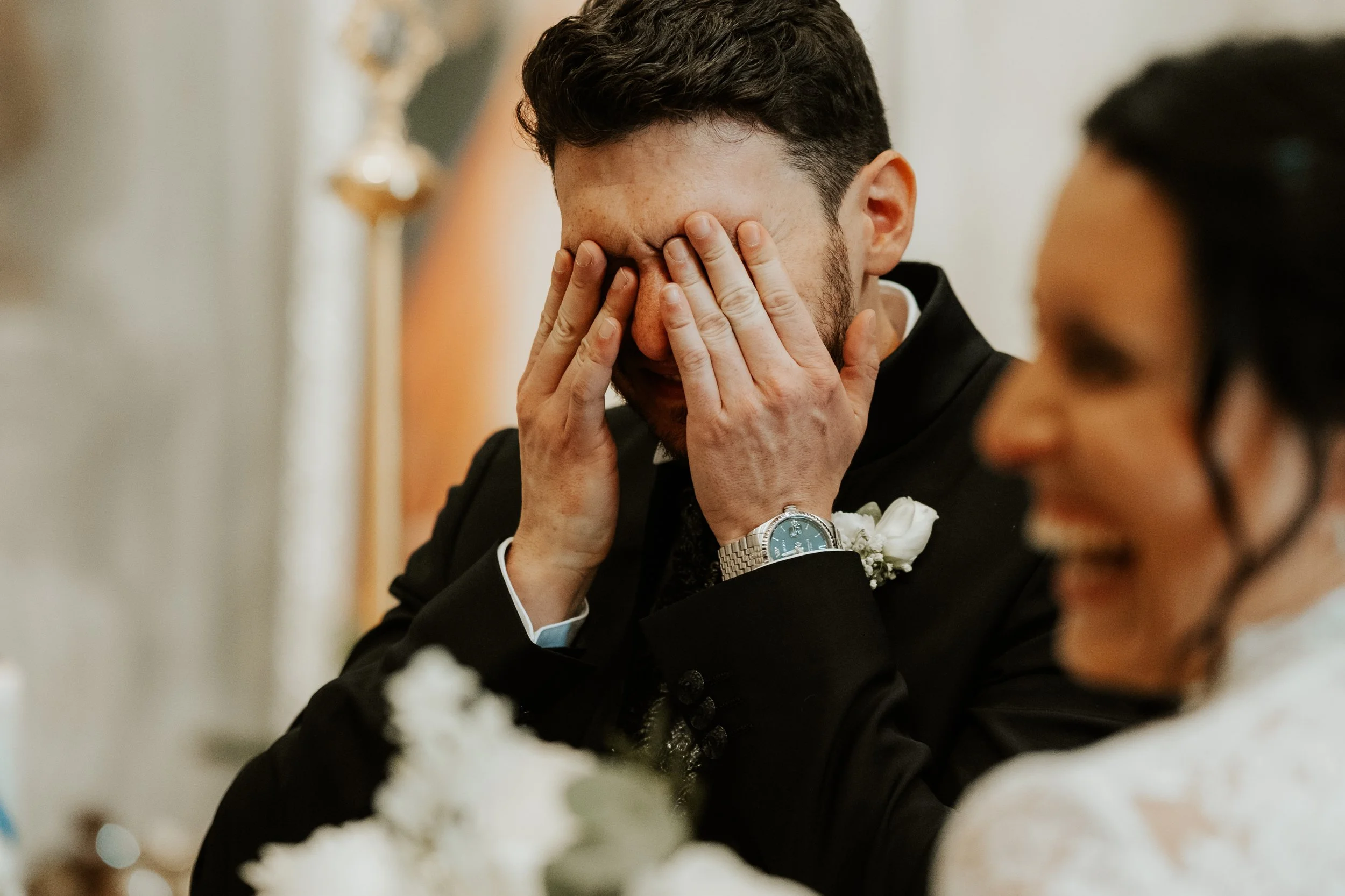 Un uomo in abito da sposo copre il viso con le mani, mentre una donna sorridente si trova accanto e all'ombra.