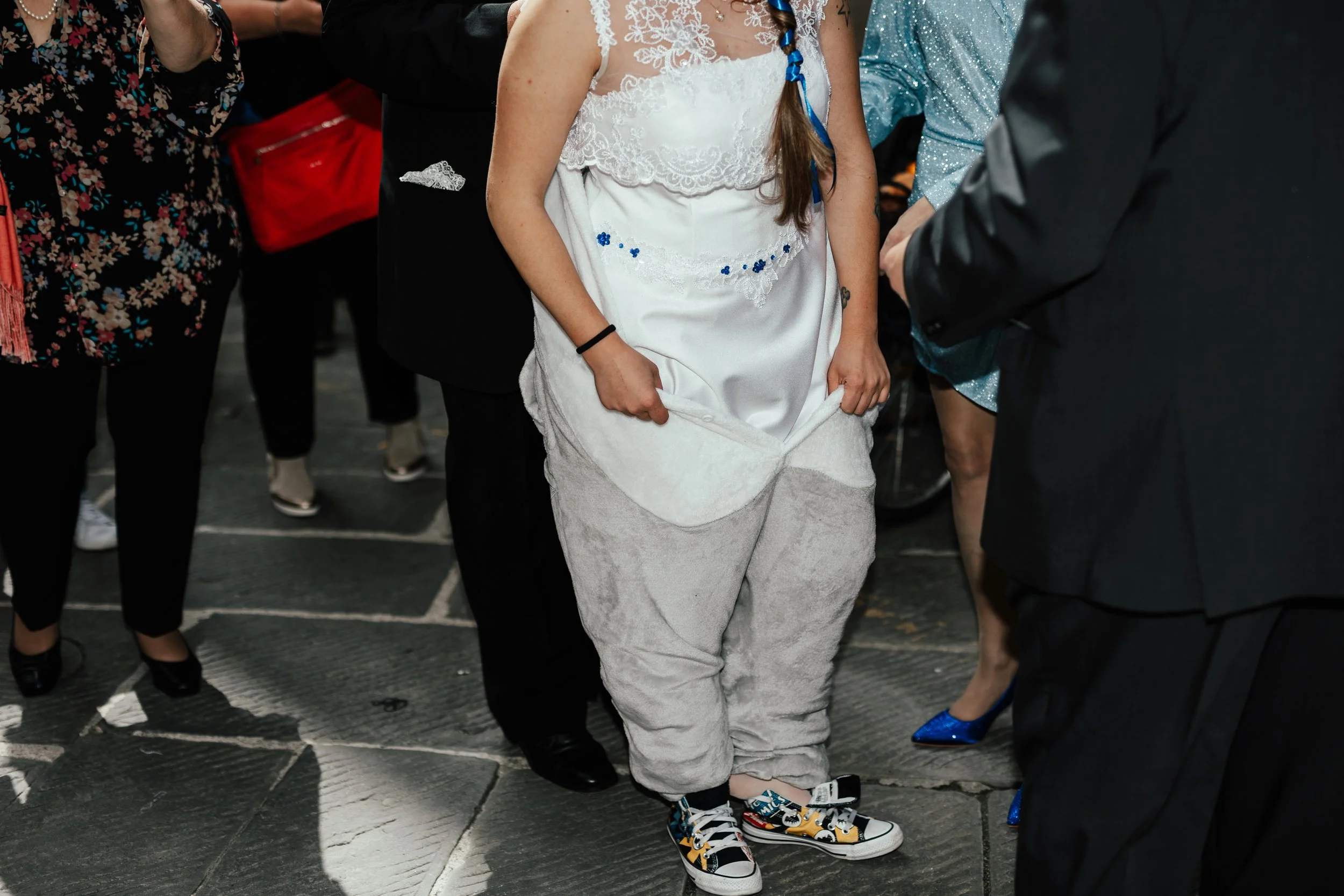 Una persona vestita con un abito bianco da sposa, con pantaloni morbidi e scarpe da ginnastica colorate, si trova tra altri individui in abiti eleganti e casual durante un evento sociale.