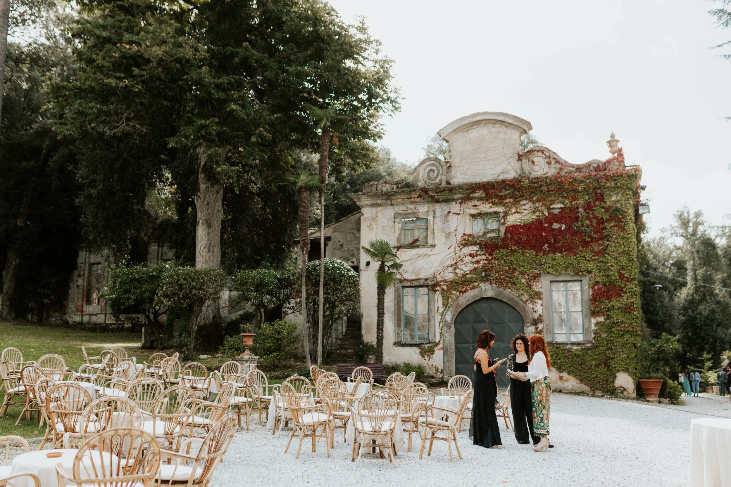 Tre donne che conversano davanti a un antico edificio con muri coperti di edera, circondato da sedie e tavoli imbanditi, in un cortile esterno. Villa di Corliano