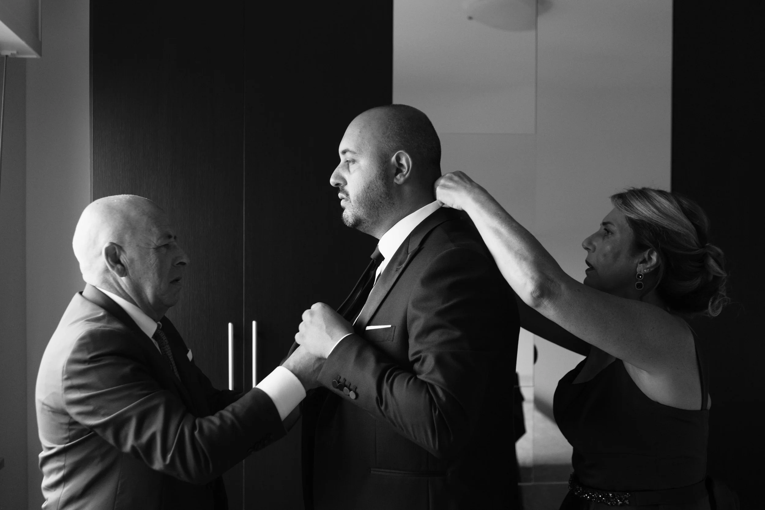 Un uomo vestito con un abito elegante viene aiutato da una donna a sistemarsi la giacca, mentre un altro uomo con un abito svolge un gesto di sostegno o approvazione. L'immagine è in bianco e nero.