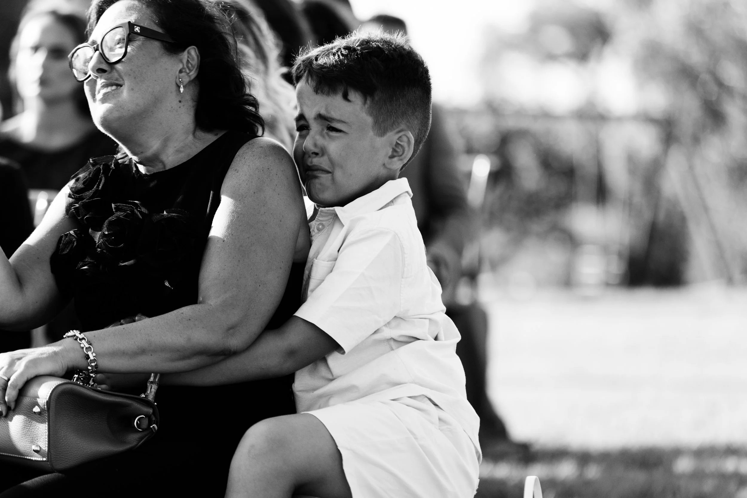 Un bambino che piange, seduto sulle ginocchia di una donna anziana, che sembra essere triste, in un ambiente all'aperto.