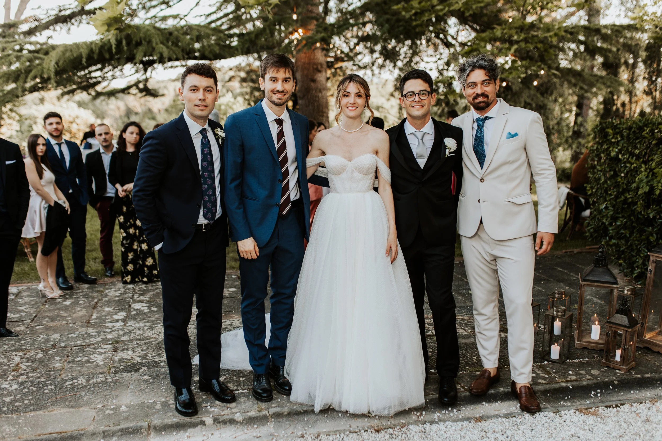 Gruppo di persone in abiti eleganti, tra cui una donna in abito da sposa bianco, al matrimonio, all'aperto con alberi sullo sfondo.