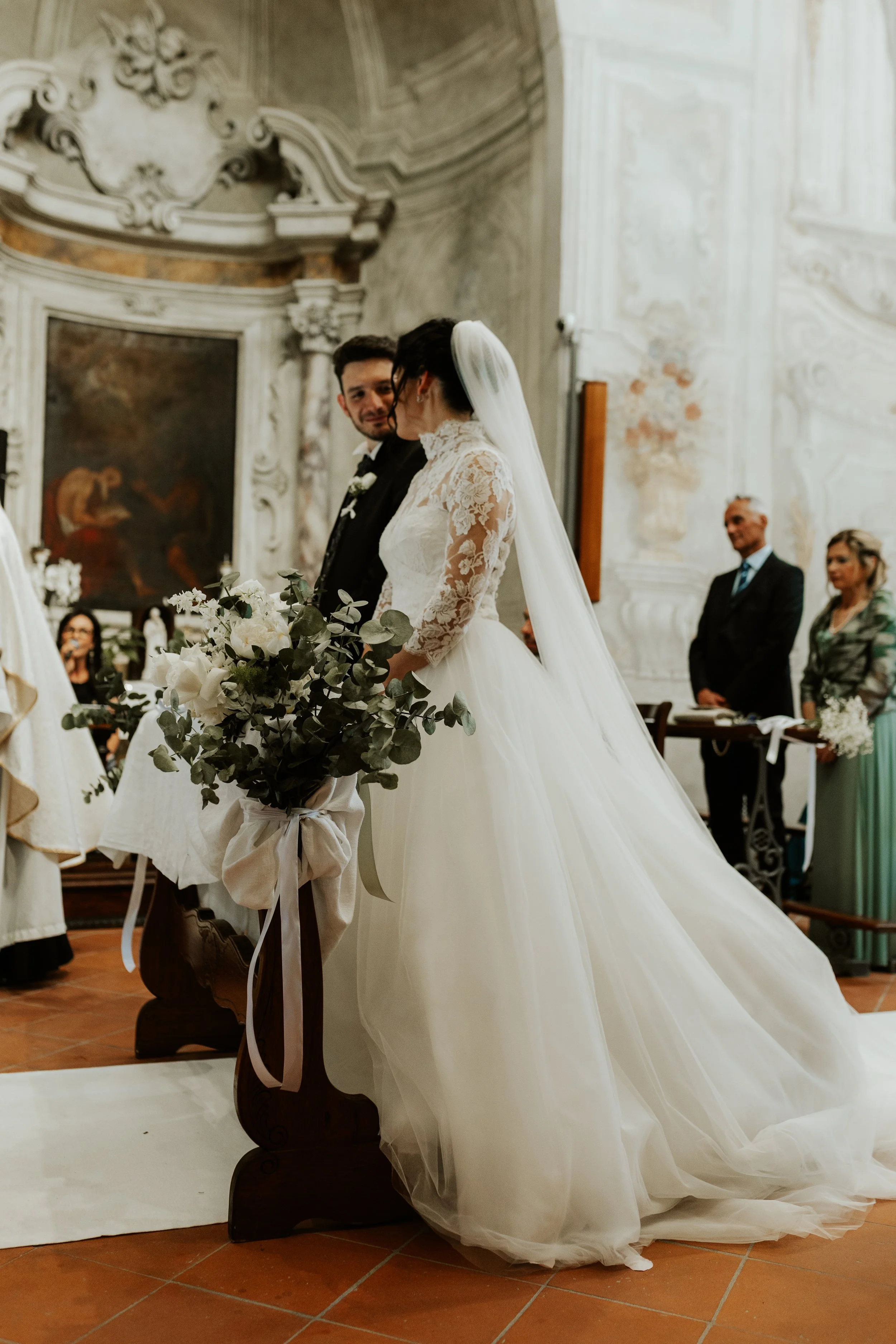 Sposi al matrimonio in chiesa con lo sposo che guarda la sposa. La sposa indossa un abito bianco con maniche in pizzo e un velo lungo, tiene un bouquet di fiori. Sullo sfondo, testimoni e ospiti che partecipano alla cerimonia.