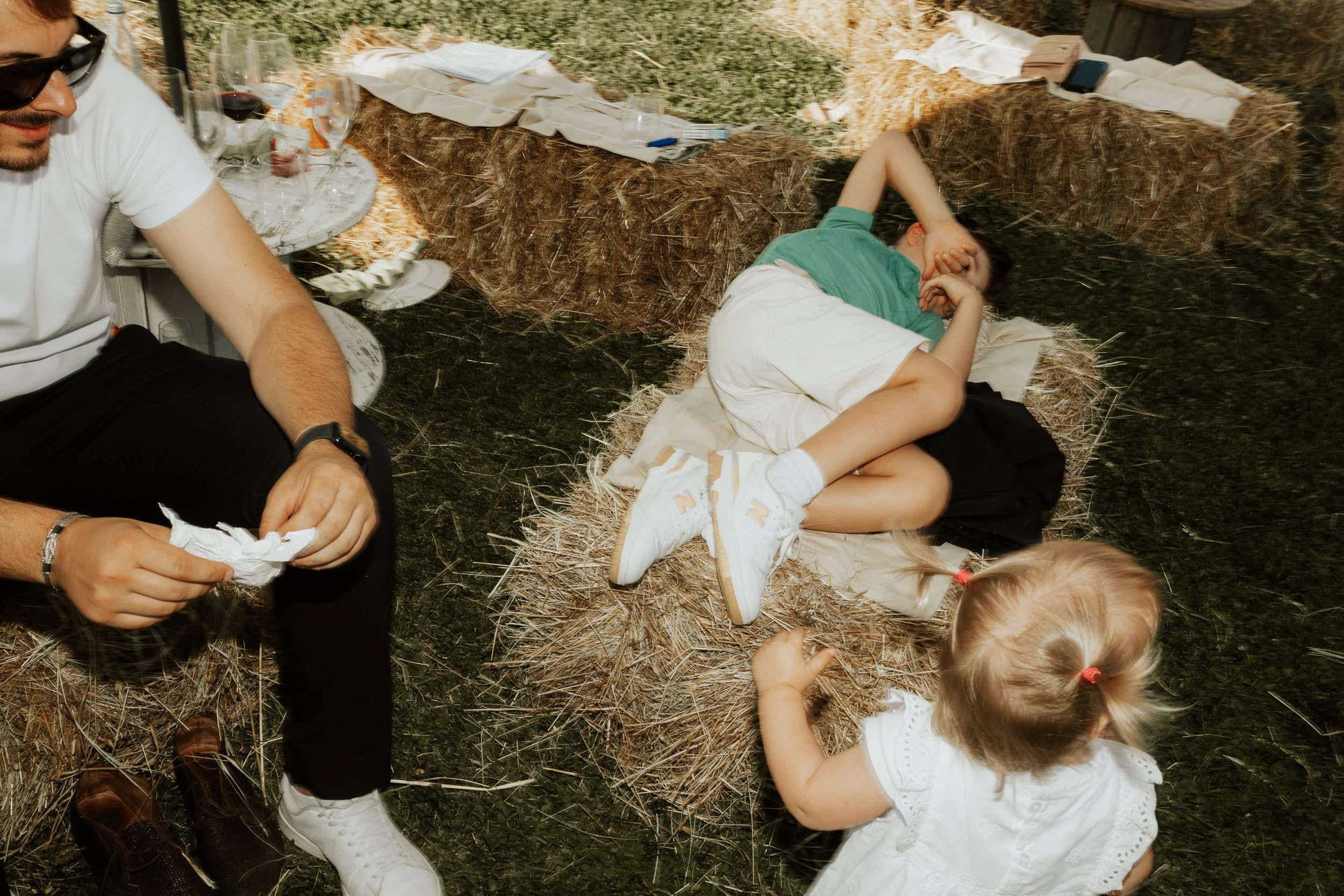 Un bambino sdraiato su un pagliericcio mentre un’altra bambina si avvicina, in un ambiente rustico con balle di paglia e un tavolo con cibo e bicchieri.