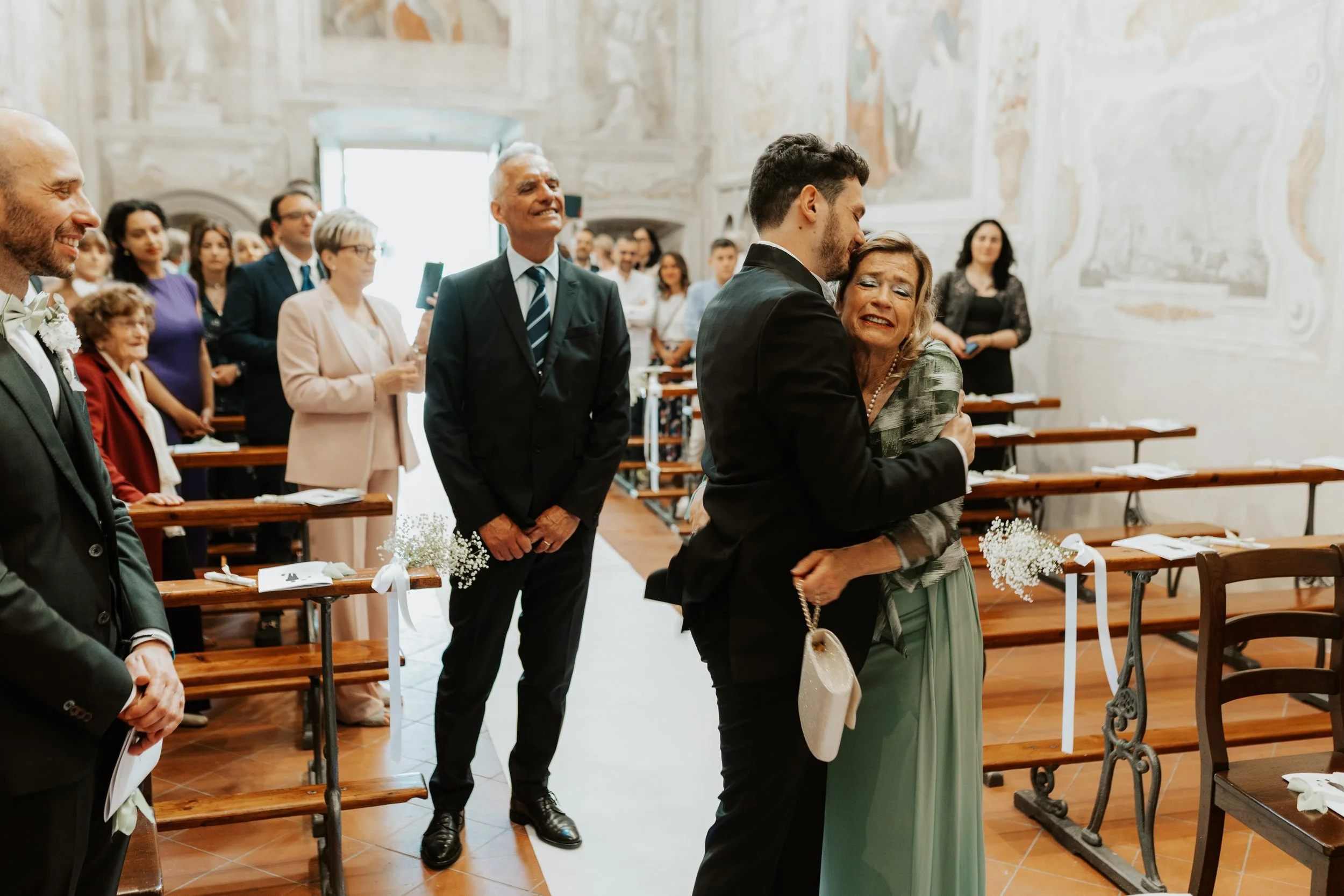 Uomo e donna si abbracciano emotivamente durante un matrimonio in una chiesa, con altri invitati sorridenti sullo sfondo.