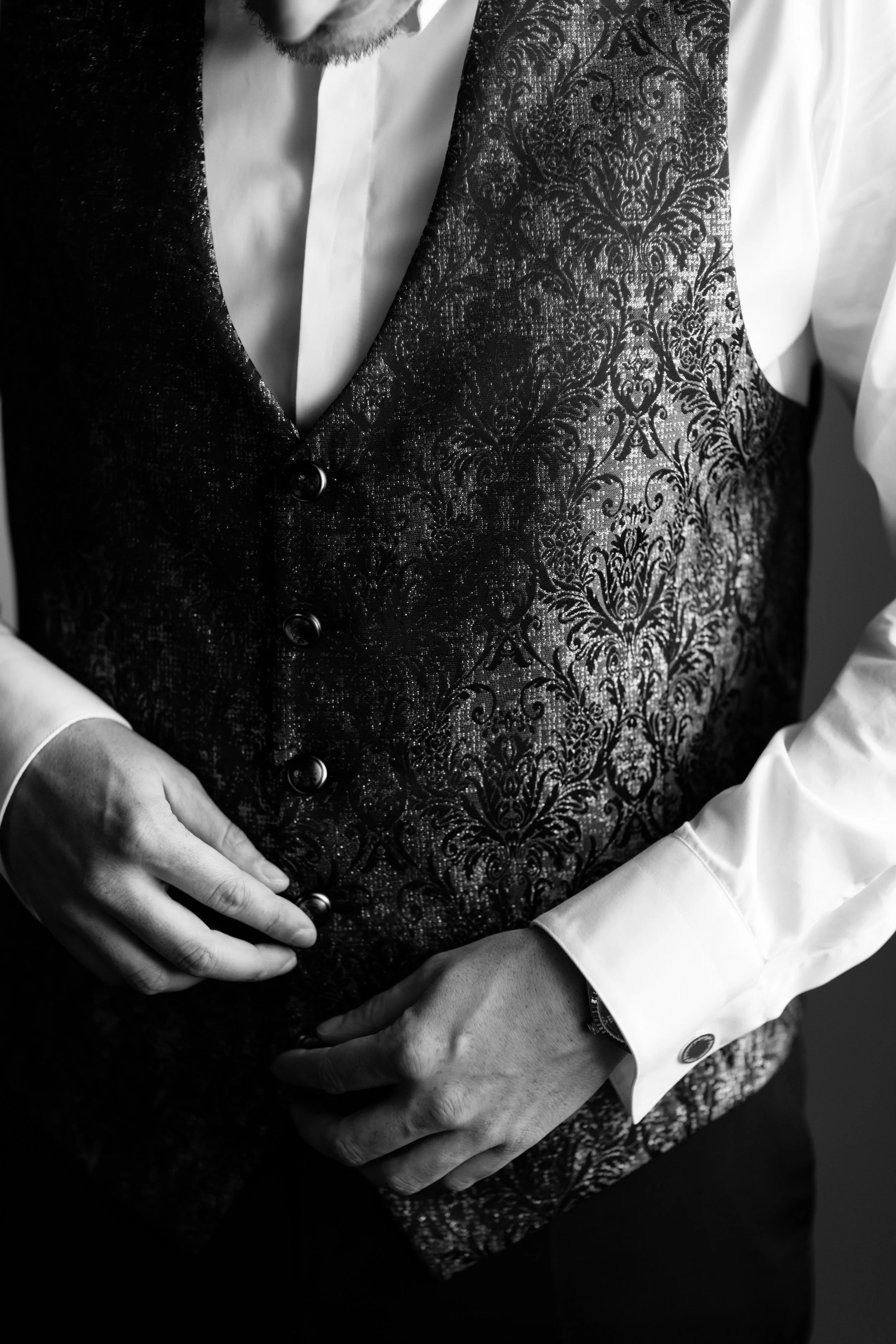 Uomo che indossa un abito nero con velluto e una camicia bianca a maniche lunghe, con mani che reggono il vestito davanti.