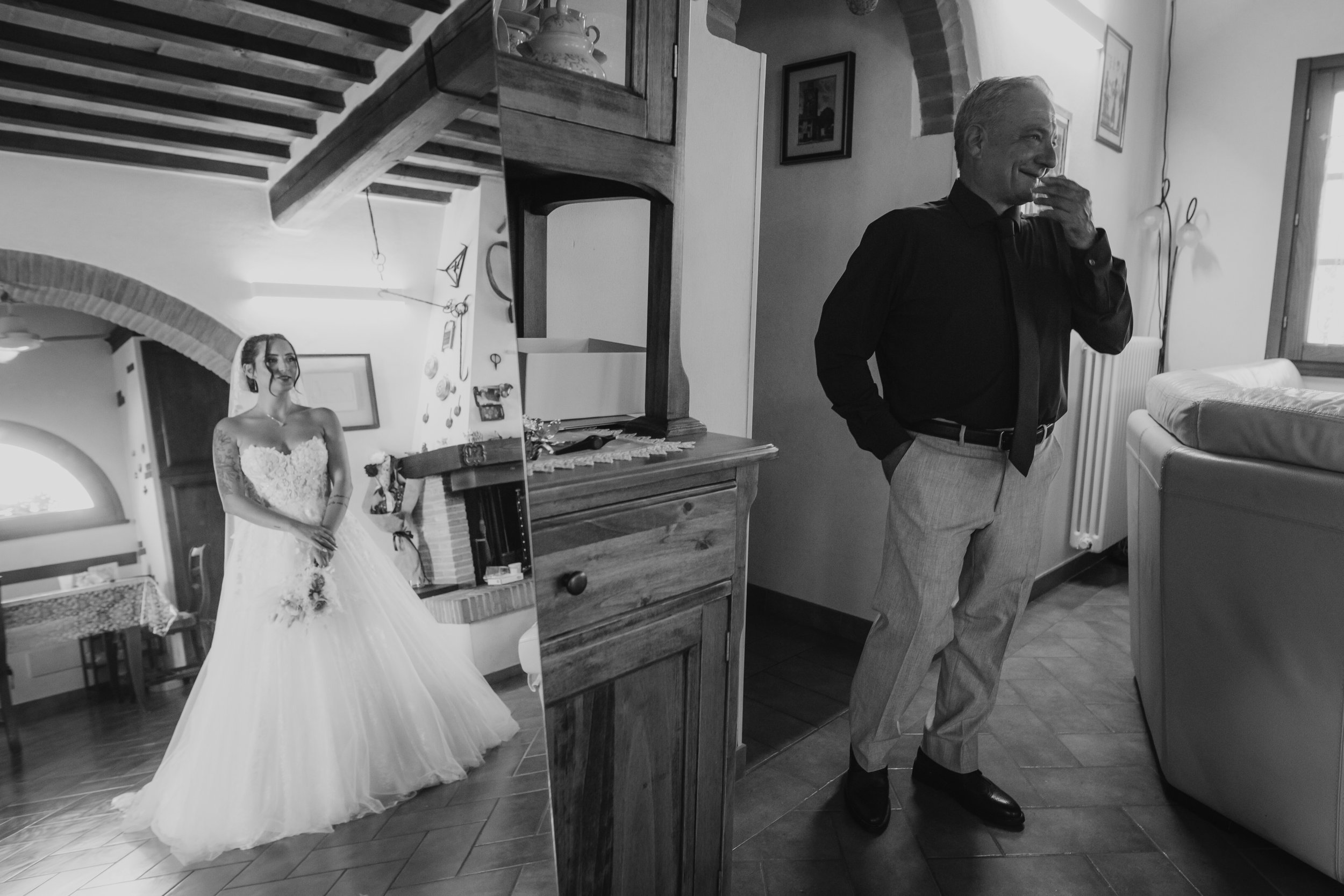 Ritratto in bianco e nero di un uomo anziano vestito in modo formale, sorridente, che si nasconde la bocca con una mano, in una stanza con pareti chiare e arredamenti classici. Sullo sfondo, un grande specchio riflette una donna in abito da sposa, so