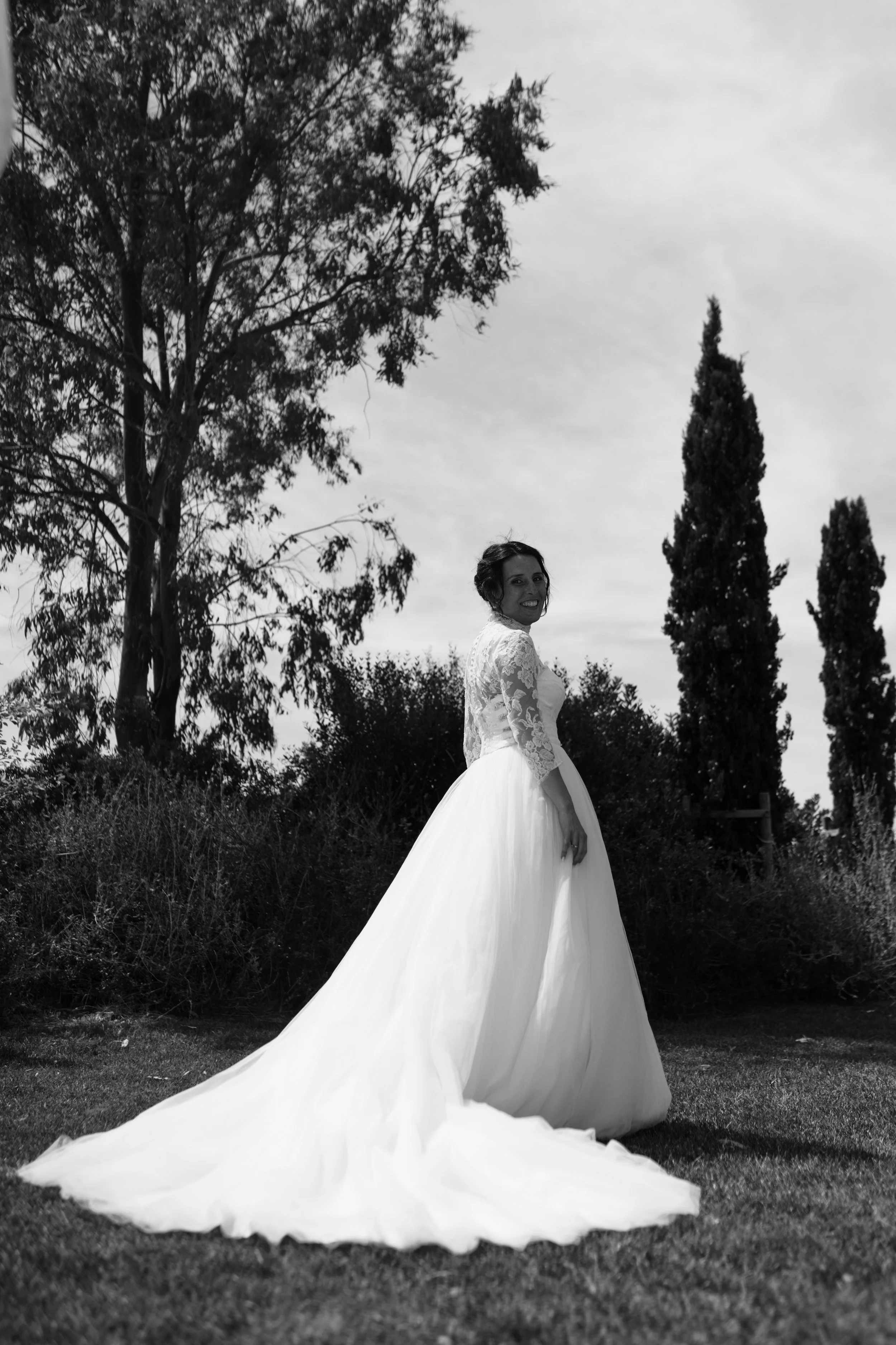 Una donna in abito da sposa bianco all'aperto, circondata da alberi e arbusti, sotto un cielo nuvoloso, in una fotografia in bianco e nero.