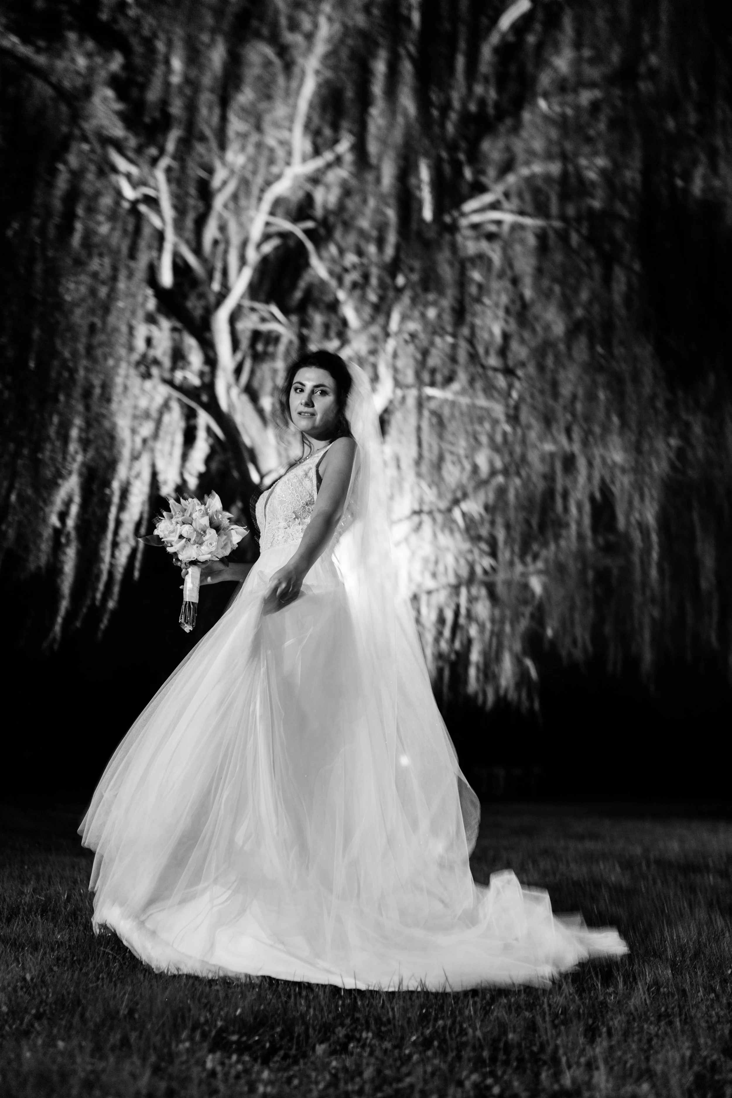 Sposa con abito lungo e bouquet di fiori, fotografata di notte con sfondo di alberi illuminati, in bianco e nero.