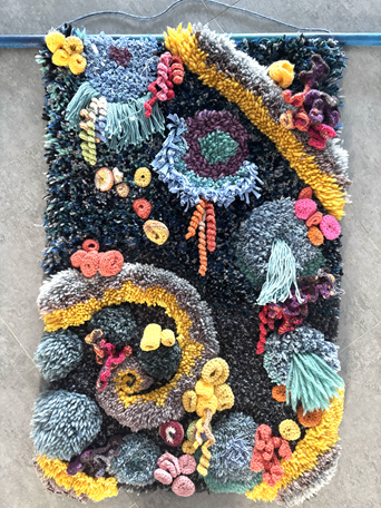 Colorful, textured handmade rug with various yarn knots, pom-poms, and tassels in bright colors including yellow, blue, pink, purple, and red, arranged in swirling and circular patterns.