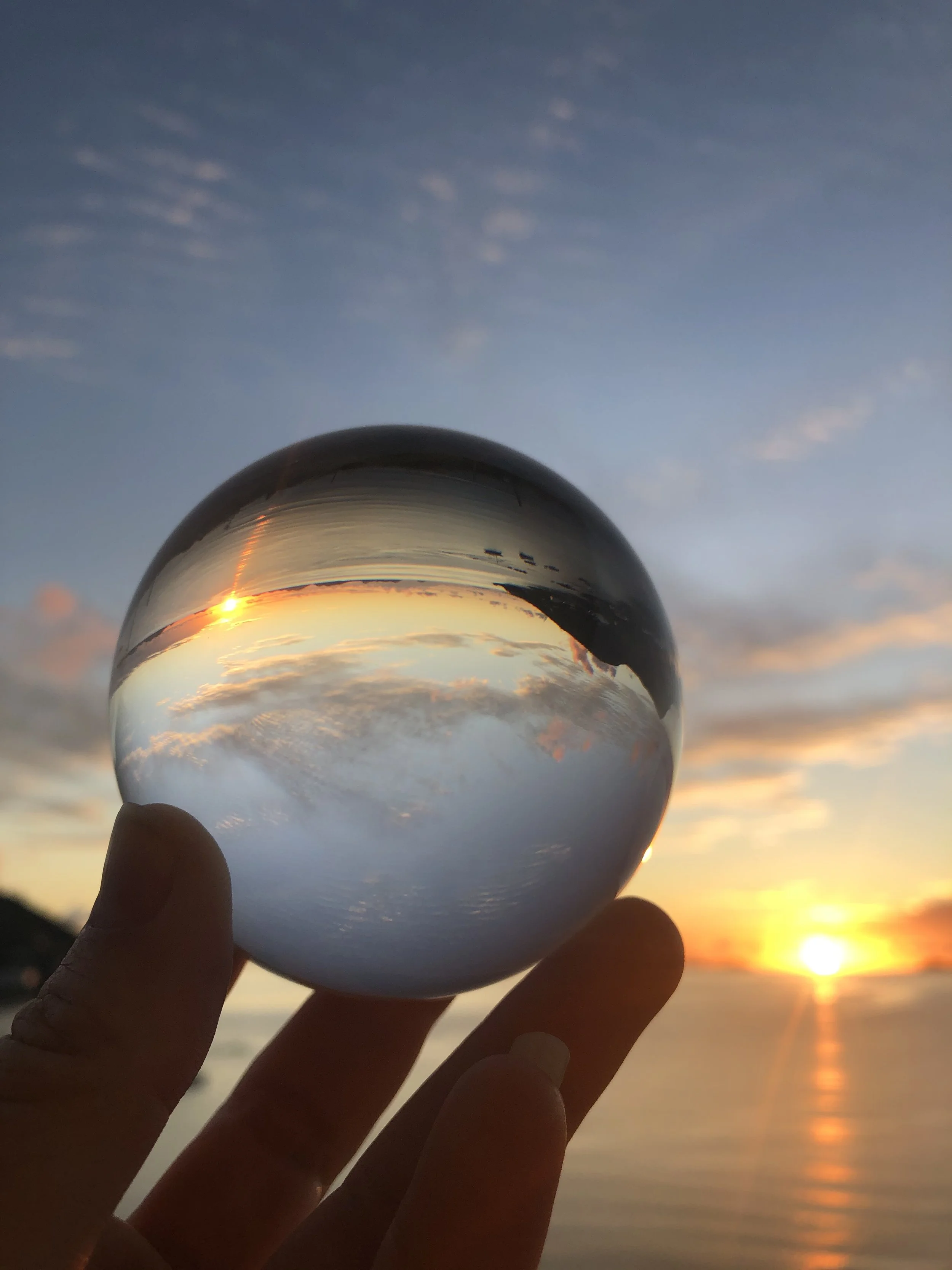 A hand holding a glass crystal ball reflecting a sunset over a body of water, with the British Virgi Islands horizon visible in the background. It is a metaphor for reframing and a hopeful, happy and healthy future