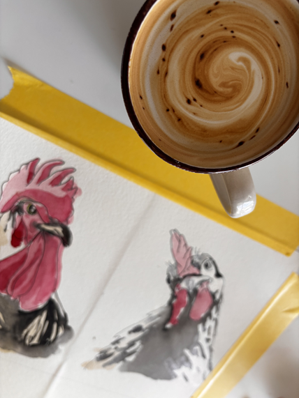 A coffee cup with swirled caramel or milk on top, and two watercolor paintings of chickens on a white surface with a yellow edge.