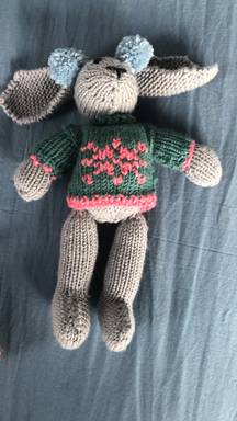 Knitted stuffed animal resembling a mouse wearing a green sweater with pink hearts and gray limbs, lying on a blue fabric surface.
