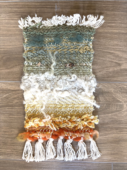 A colorful woven rug or textile piece with fringed edges, featuring various textured yarns and threads in shades of green, white, yellow, orange, and cream, laid on a wooden floor.