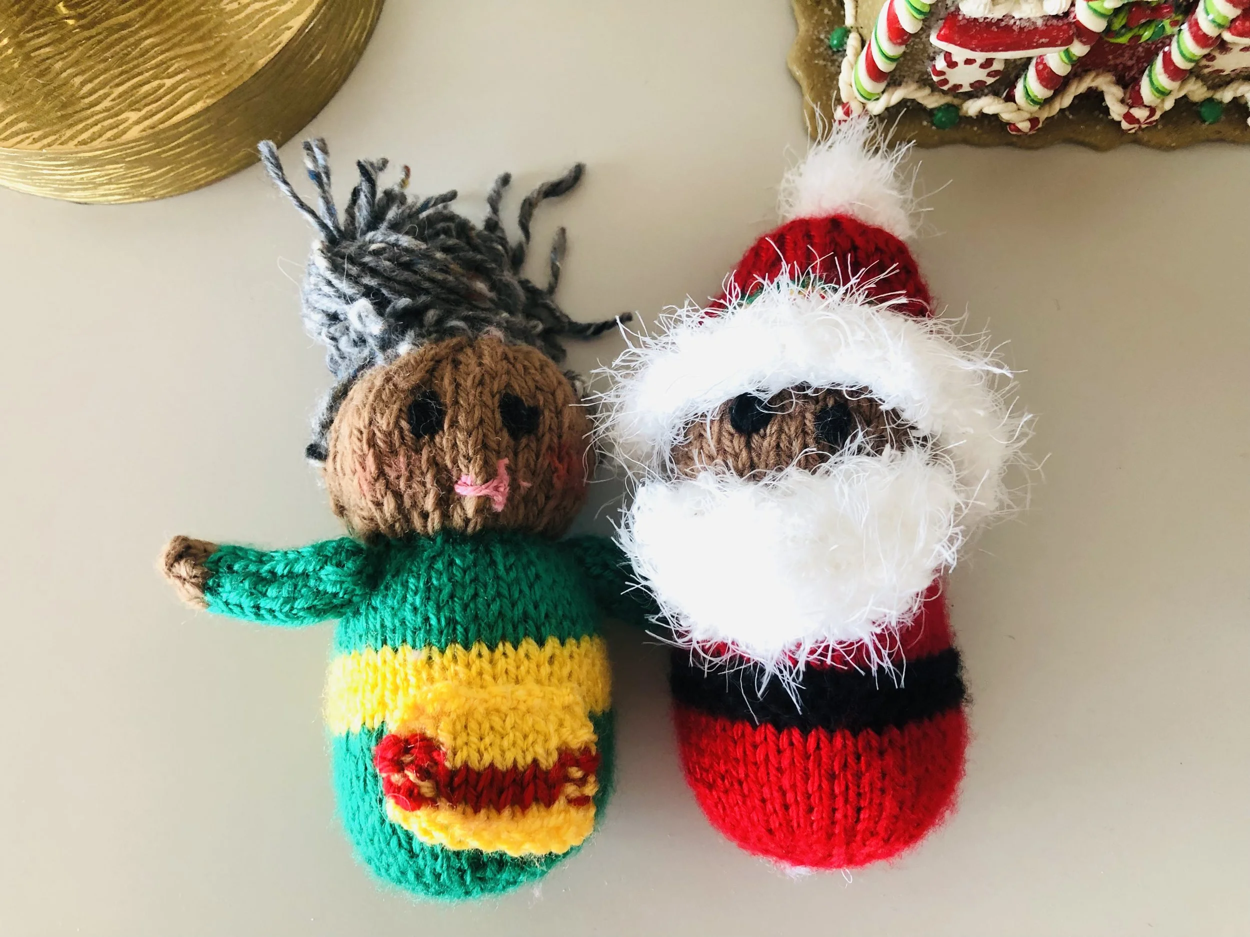 Two handmade knitted Christmas decorations: one resembles a festive elf with a brown face, pink nose, green and yellow outfit, and gray hair; the other resembles Santa Claus with a brown face, white beard, red hat, and red suit with black belt.