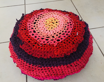 Colorful crocheted round cushion with concentric circles in pink, red, purple, and yellow, placed on a tiled floor.