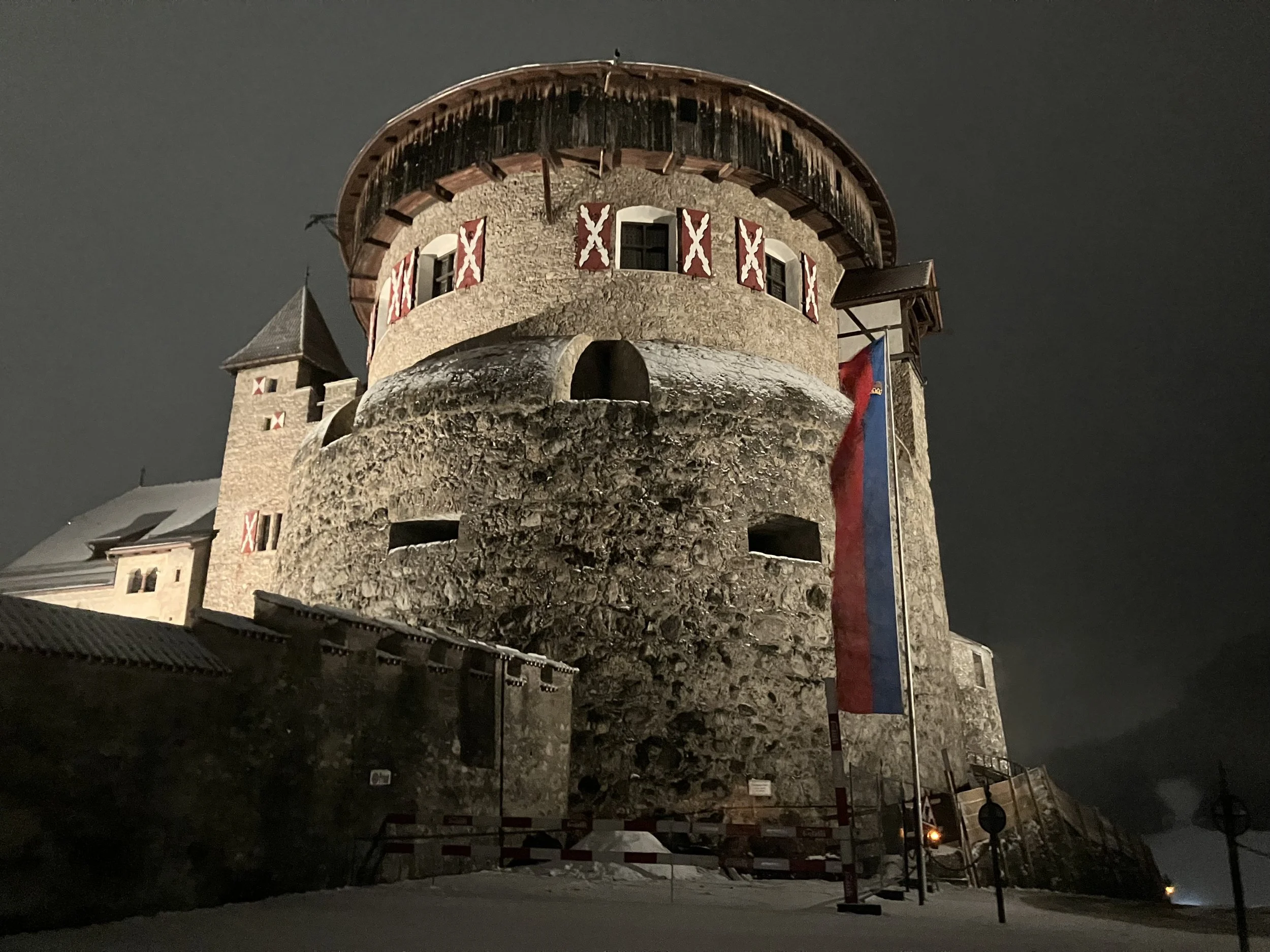 Nighttime view of a historic stone castle tower with a wooden roof, windows with red and white shutters, and a large flag hanging on the side, surrounded by snow.