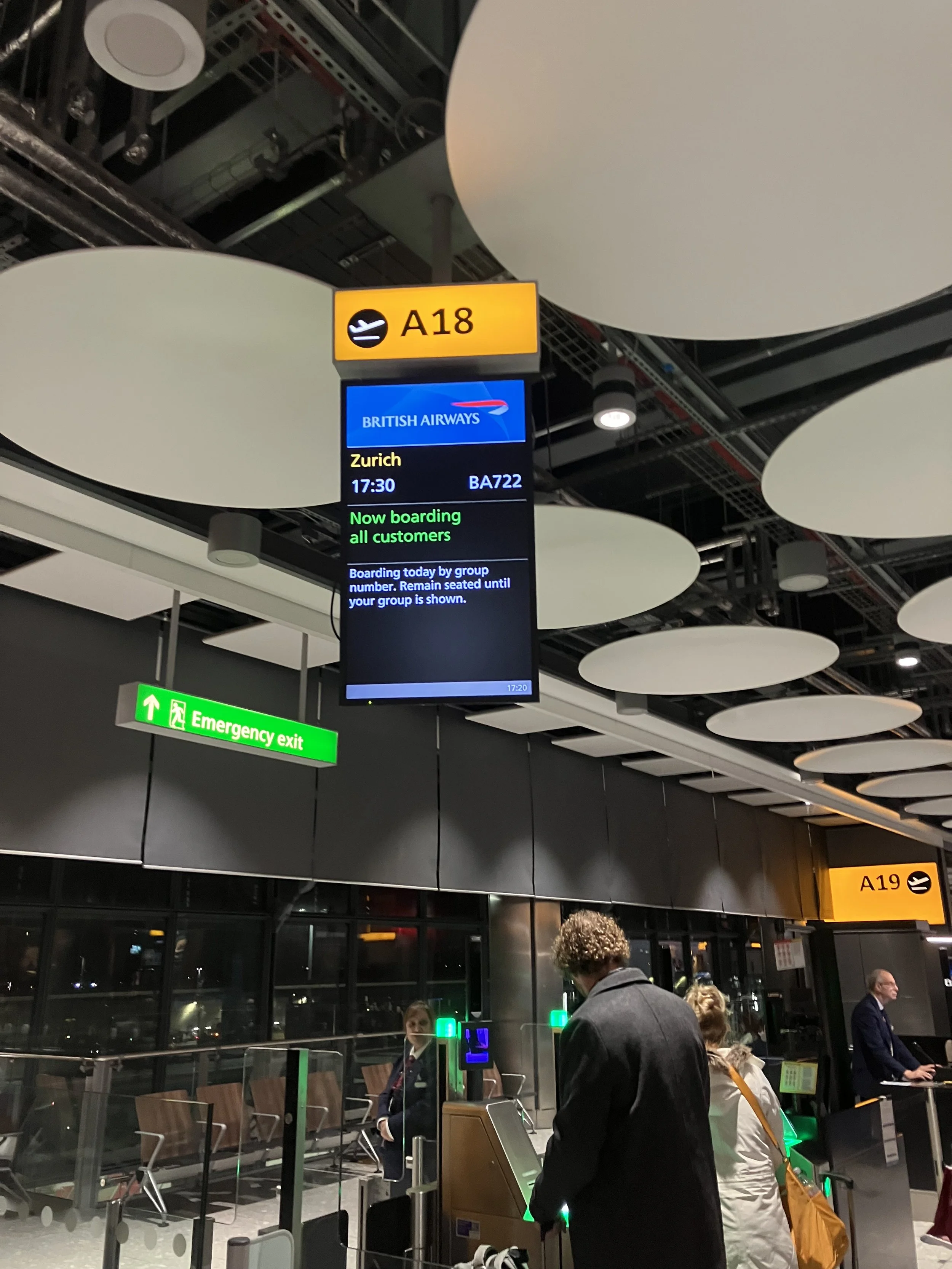 Passengers at Heathrow Airport gate A18 waiting to board a British Airways flight to Zurich, with an electronic flight information display and an emergency exit sign visible.