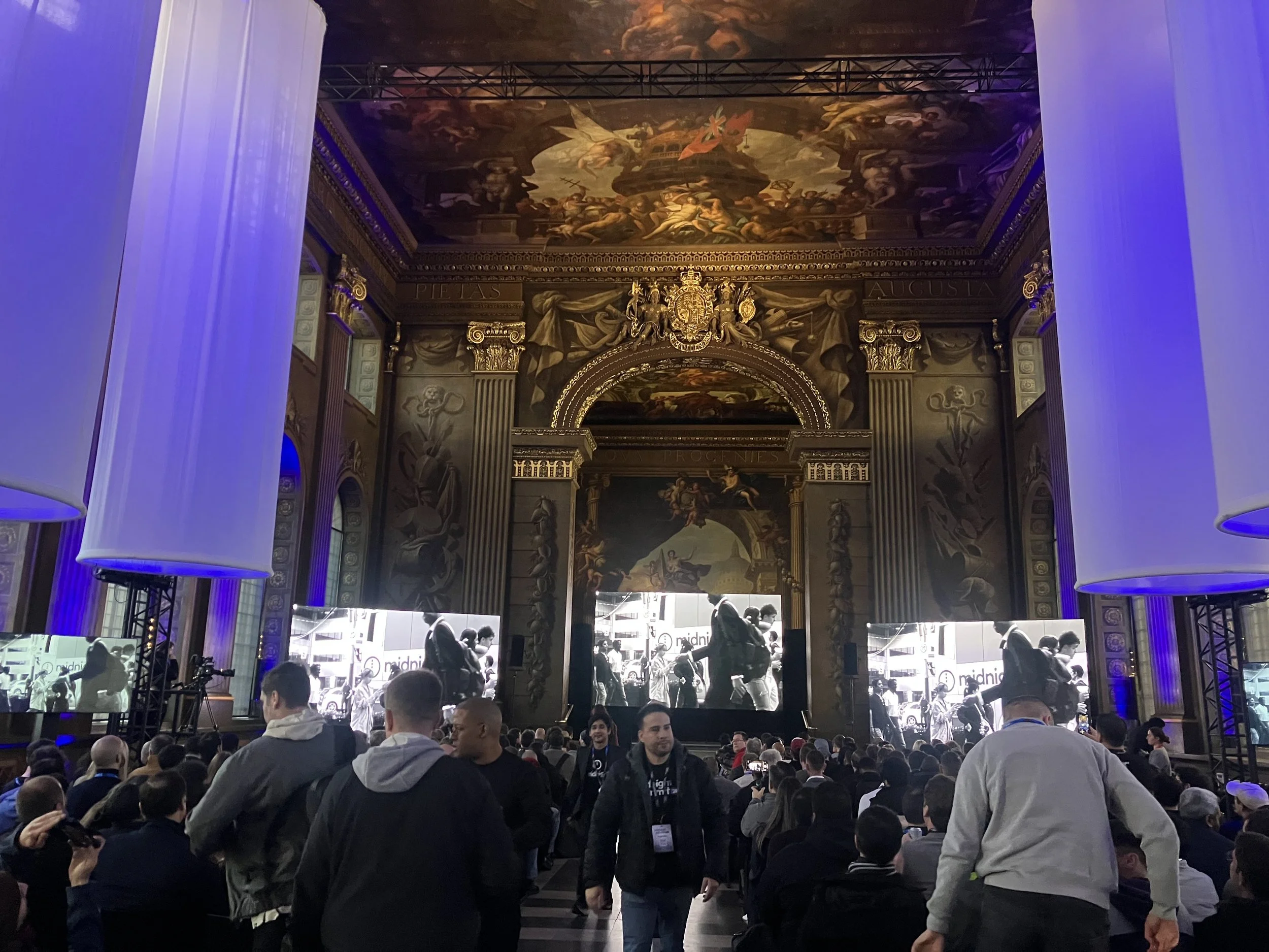 Crowd of people attending a conference or event in a grand, ornately decorated hall with large screens and lighting effects.
