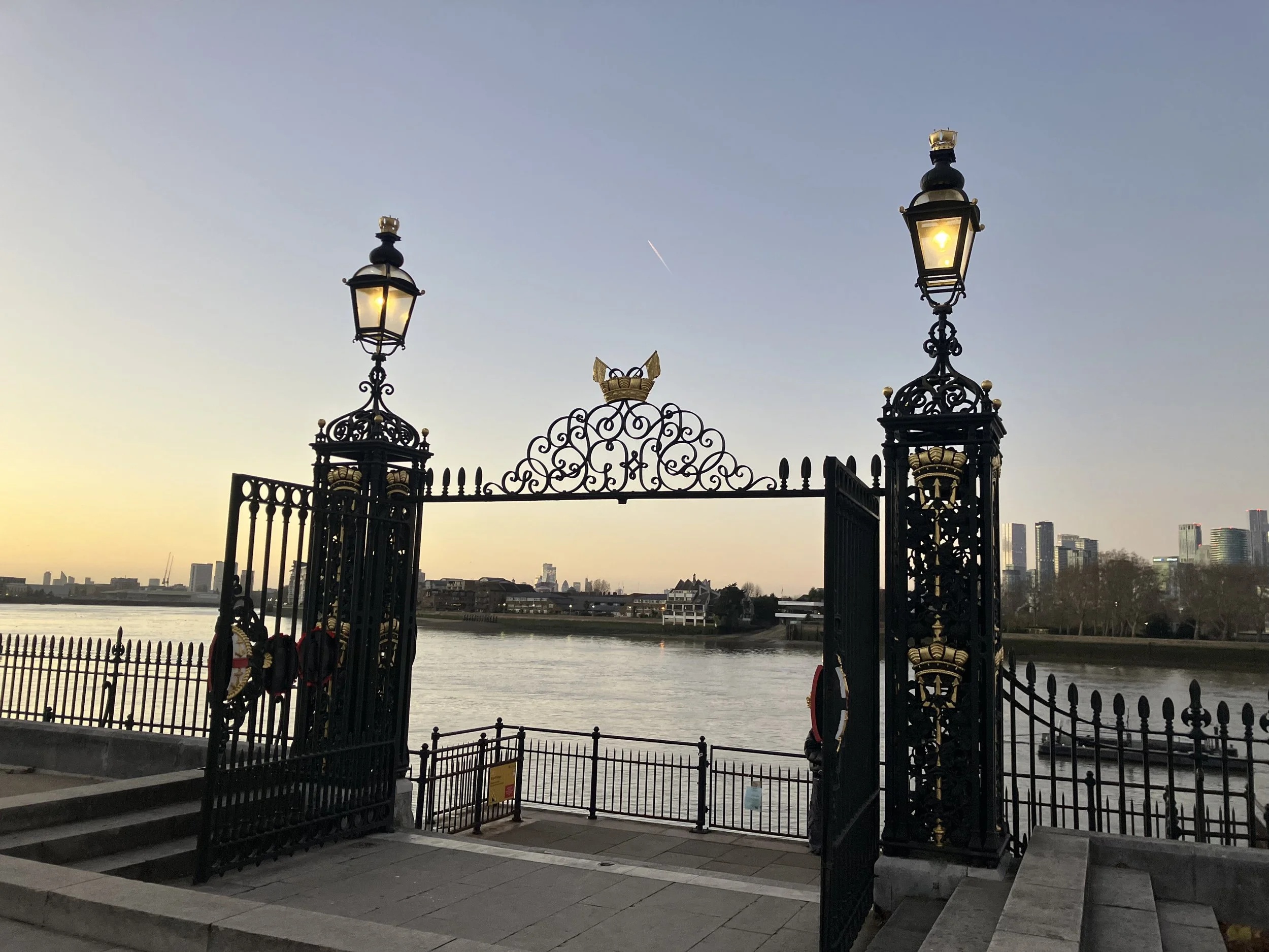 Ornate black wrought iron gates with golden accents and two glowing lamps at the top, leading to a river with a city skyline in the distance during sunset.