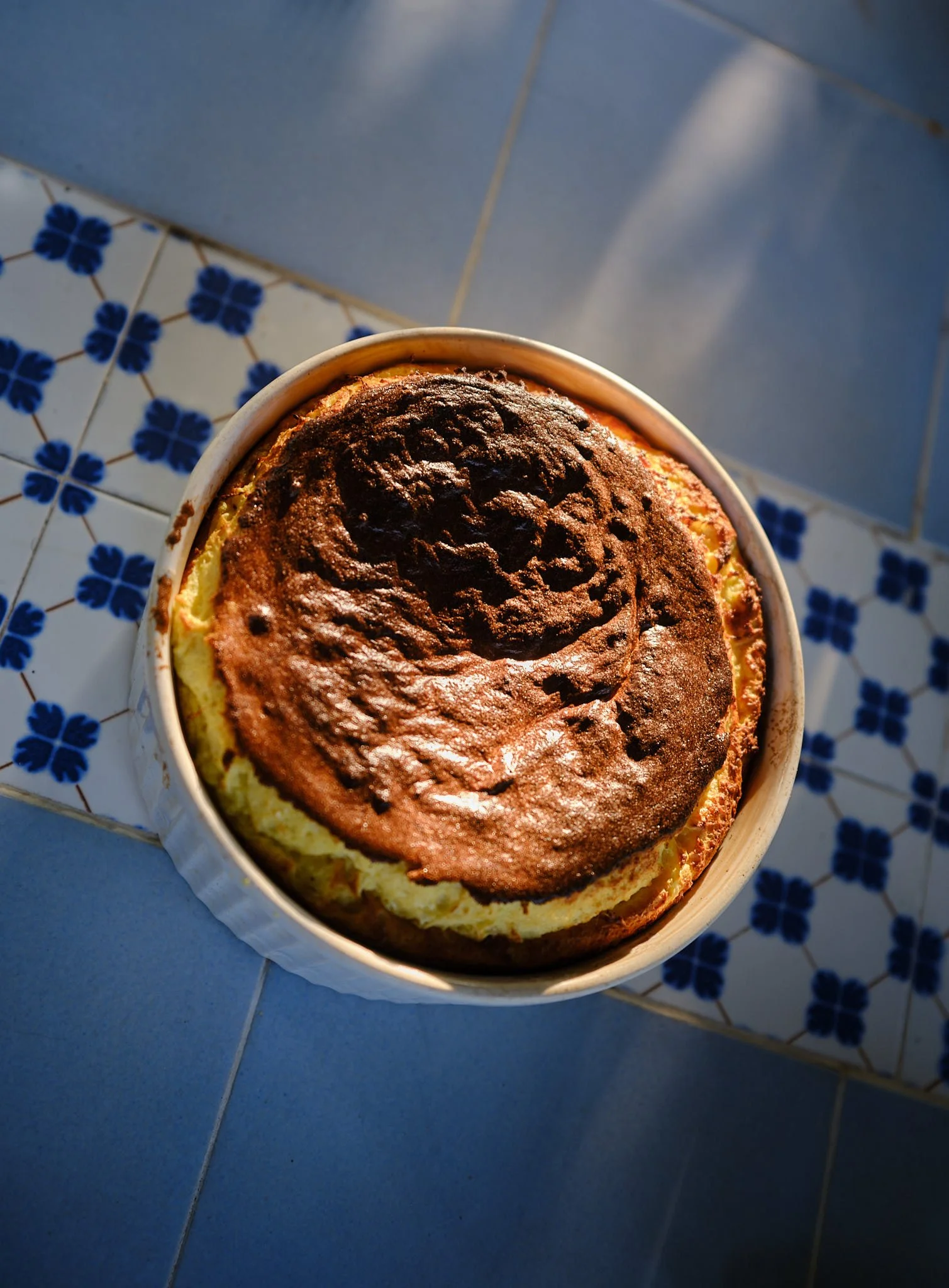 Baked cake with a burnt top in a round ceramic dish on patterned tiled floor with blue and white flower designs.