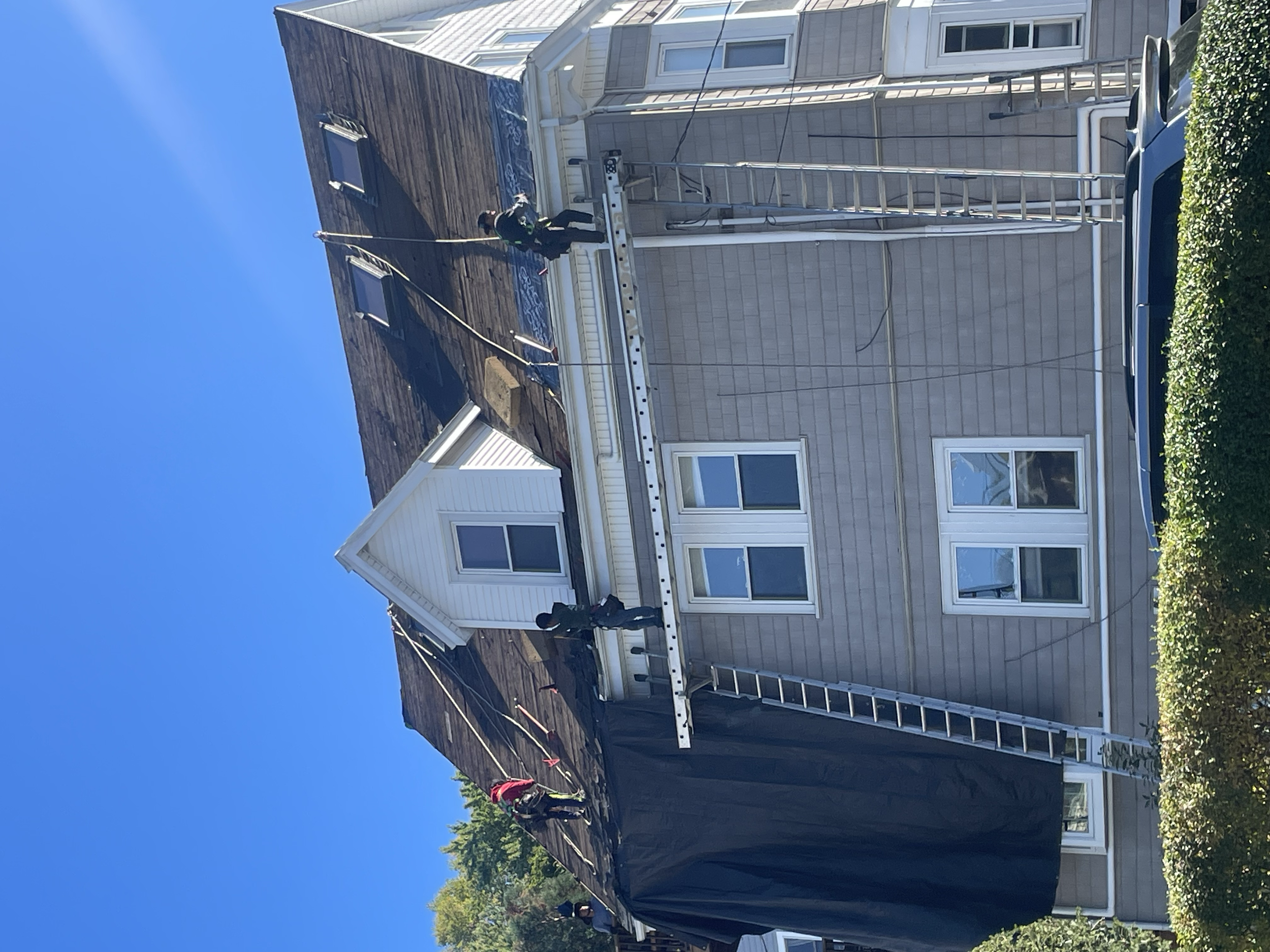 Workers repairing roof on multi-story house with ladders, tarp, and blue sky background