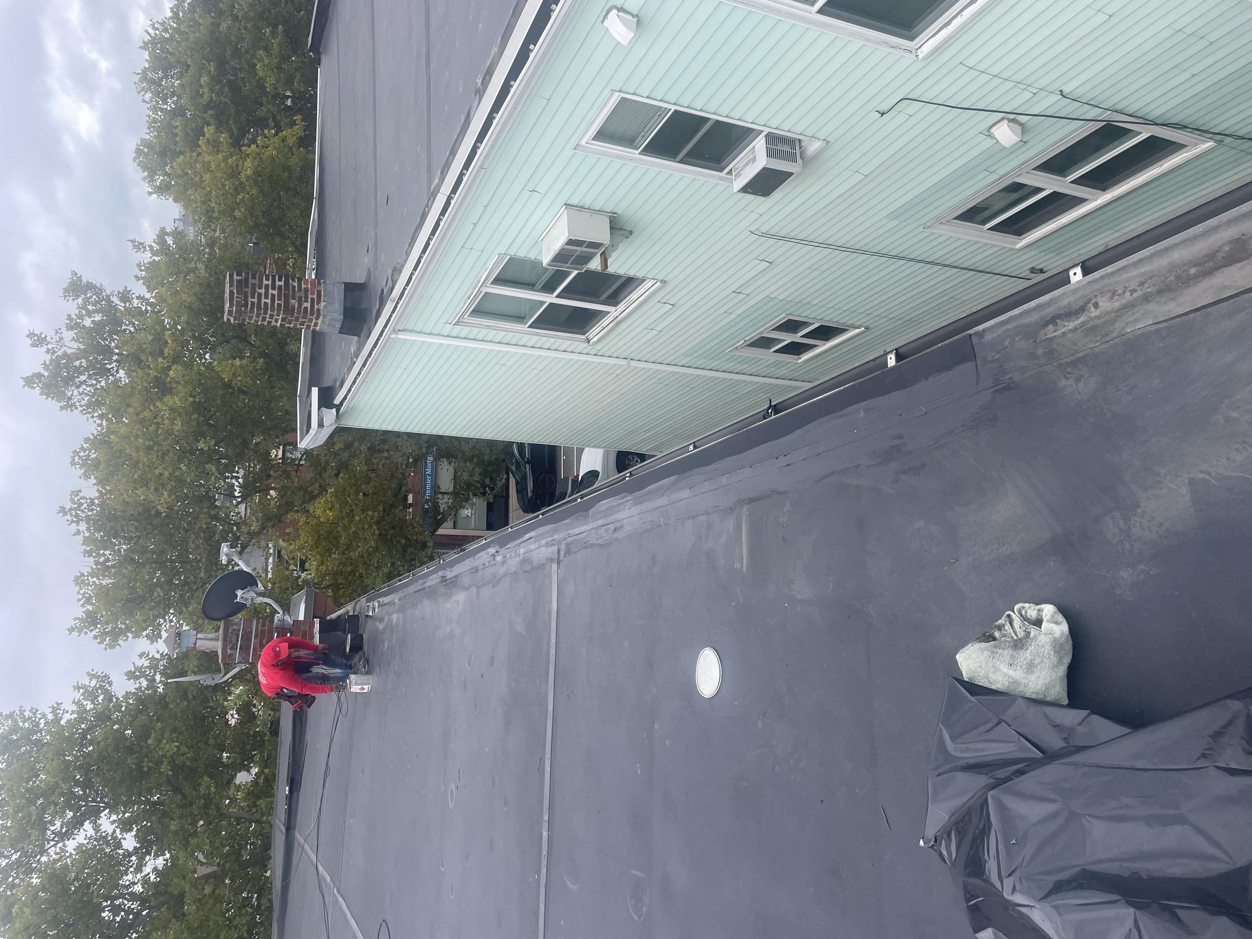 Person in red working on a flat roof with tools, near a chimney and satellite dish, next to a building with green siding and windows.
