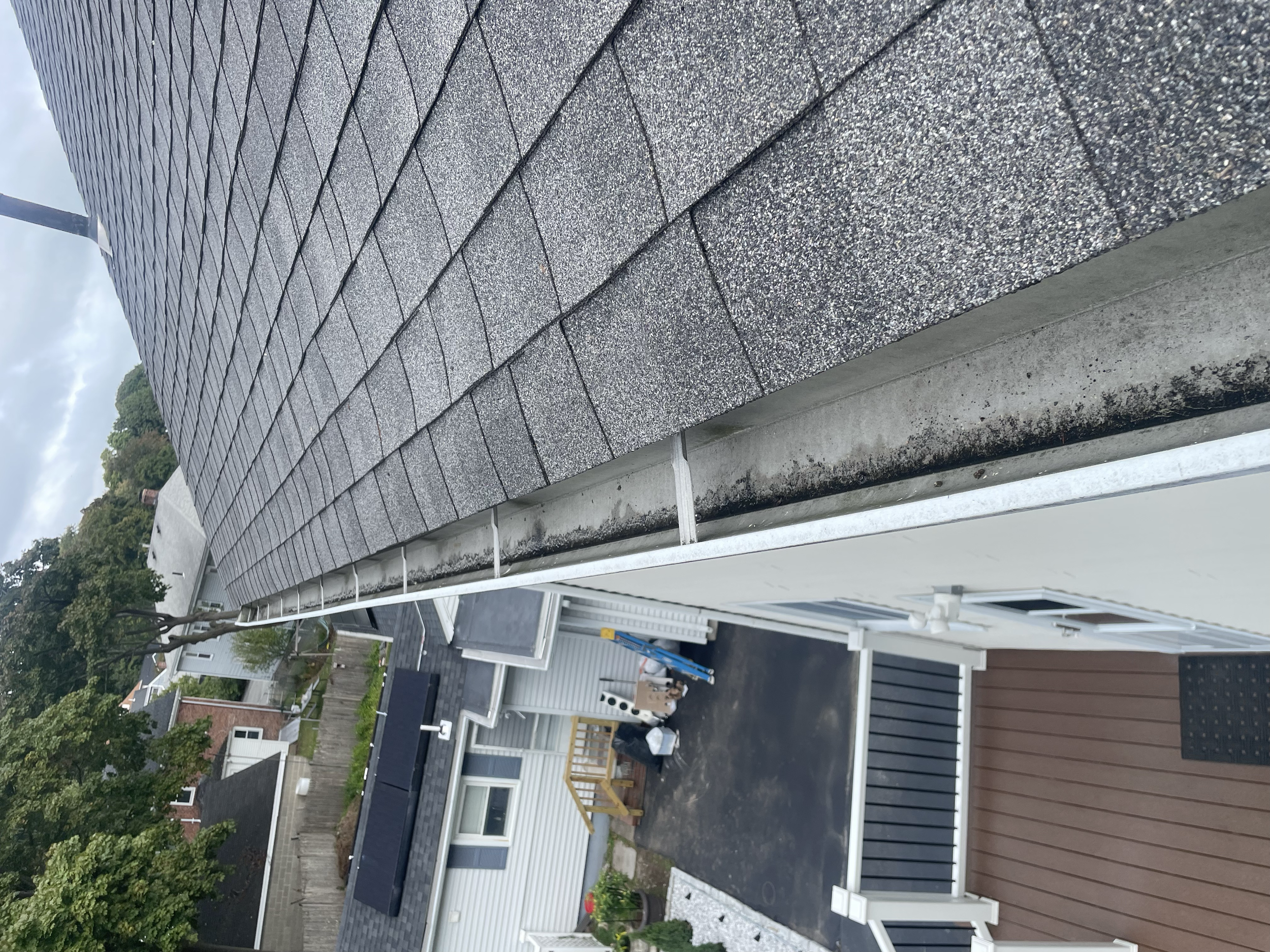 Close-up view of a house's roof with asphalt shingles and a gutter in need of cleaning, overlooking a driveway and neighboring houses.
