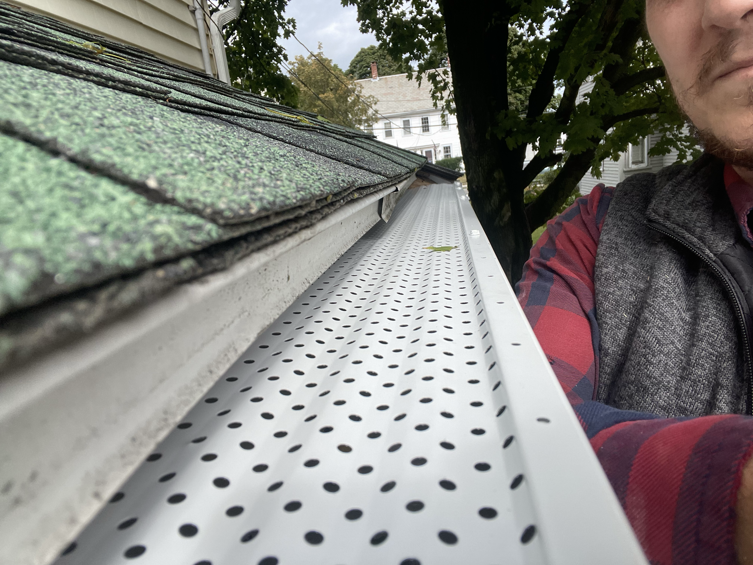 Close-up of a house roof with a gutter and gutter guard, showing green asphalt shingles on the roof. A person in a plaid shirt and vest, partially visible on the side, is present. Trees and a building in the background.