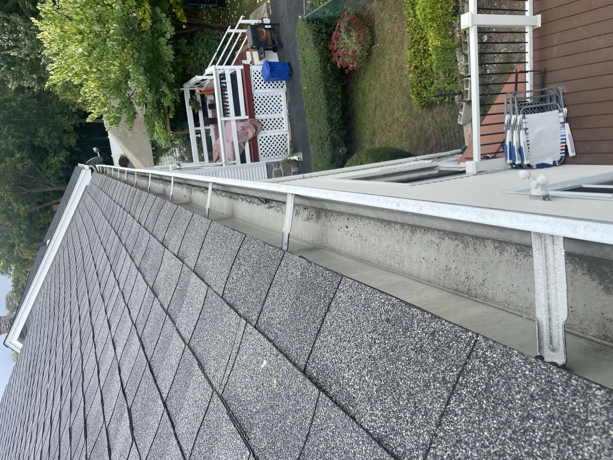 View of a residential roof with asphalt shingles, adjacent to a gutter system. Below is a deck area with outdoor furniture, surrounded by greenery.
