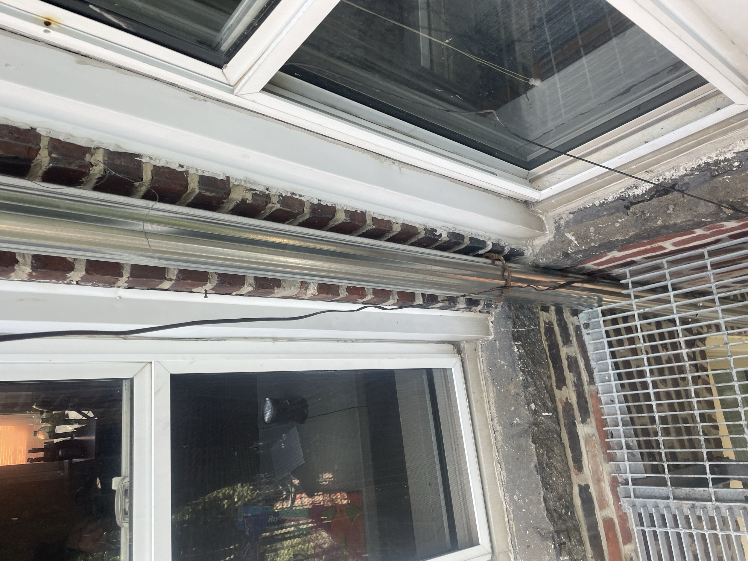 Exterior view of a building corner with a window, brick wall, and metal pipes running parallel to the structure. A metal grate covers a small vent or opening, and there are wires visible along the pipes.