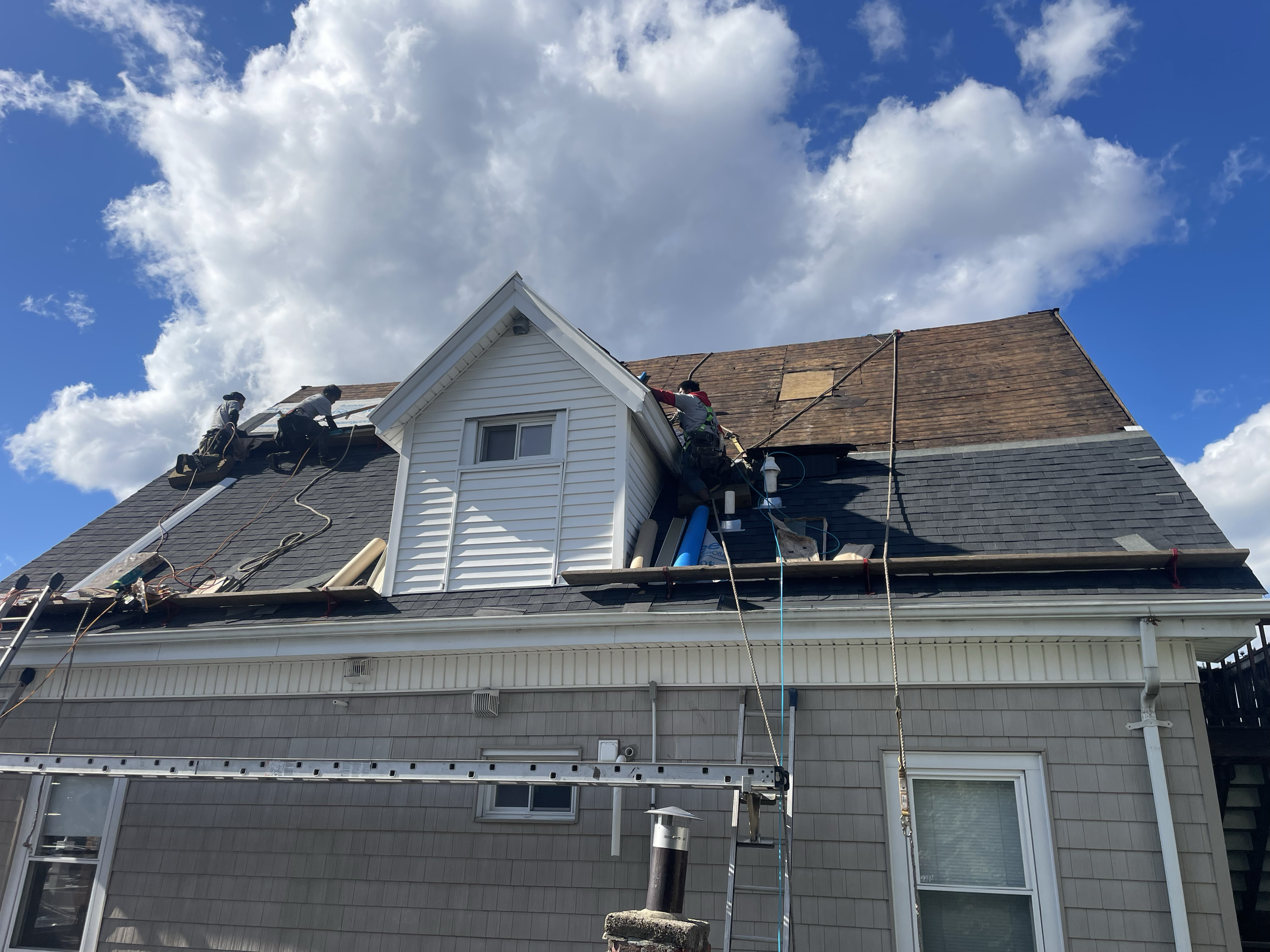 Roofers working on a house roof under a cloudy sky, using ladders and ropes, partially replacing shingles.