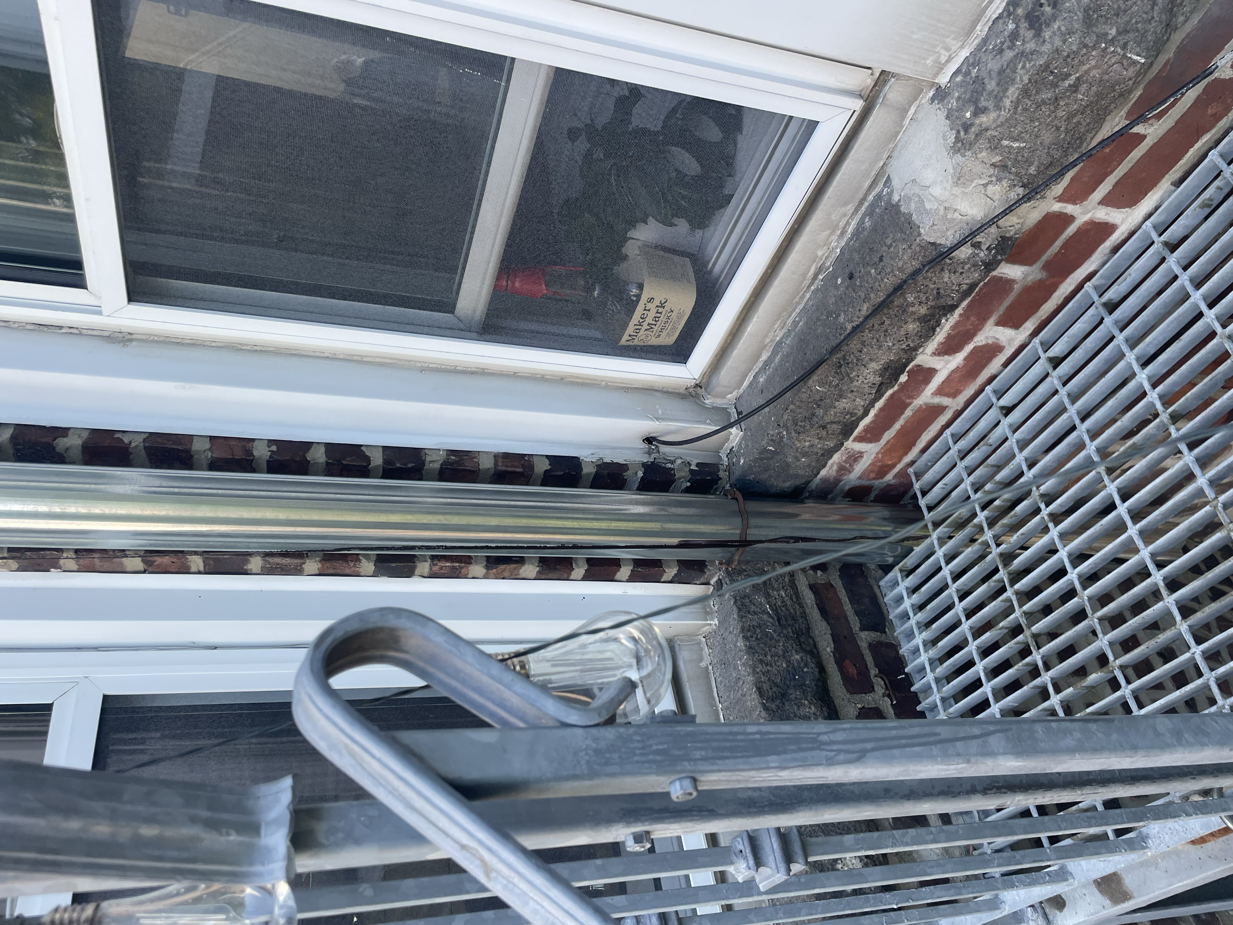 Close-up of a window with a metal grating and electrical wiring nearby. The window is surrounded by a brick wall and partially obscured by metal and glass bars.