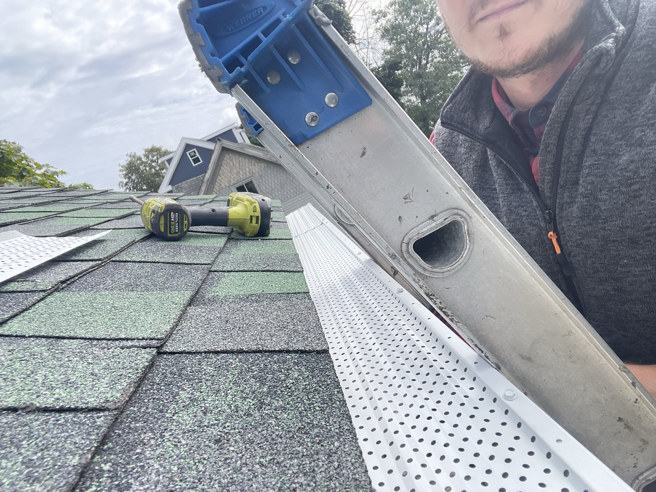 Person on roof with ladder, installing gutter guards; drill and perforated metal sheet visible.