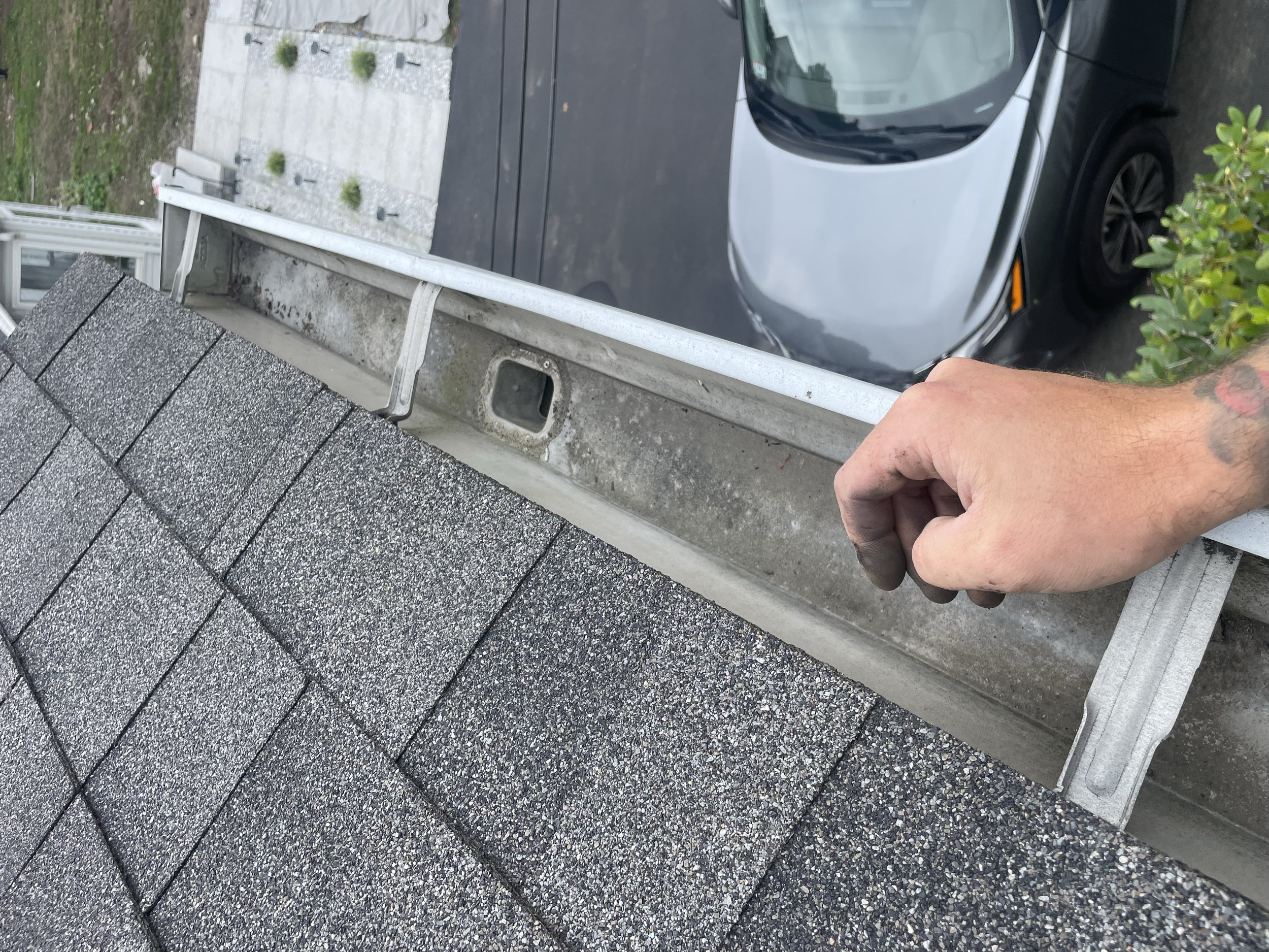 A hand inspecting an asphalt shingle roof and gutter, with a driveway and parked car below.