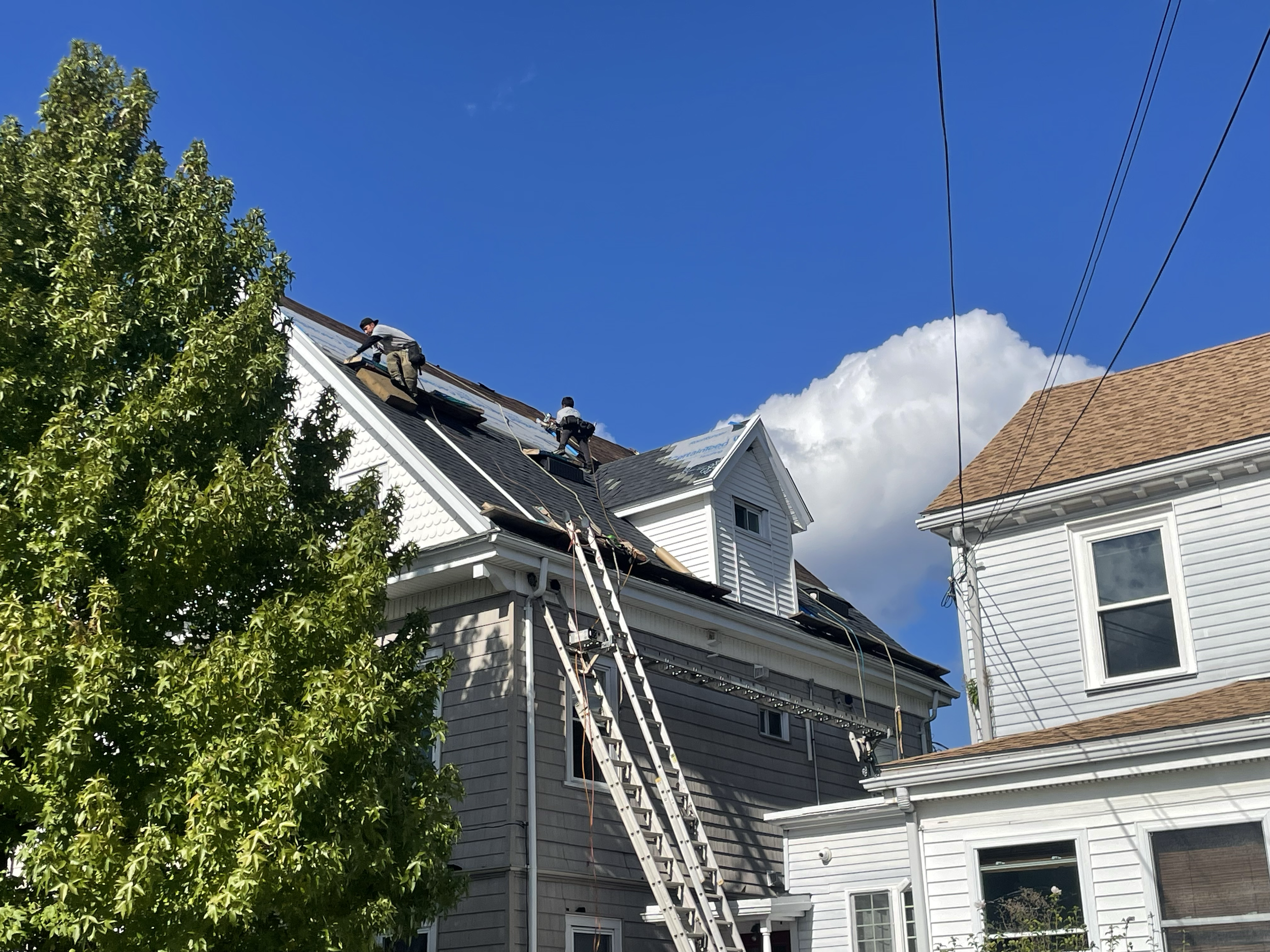 Roofers working on a steep roof of a gray house with two ladders, surrounded by a blue sky and partial view of a white house on the right.
