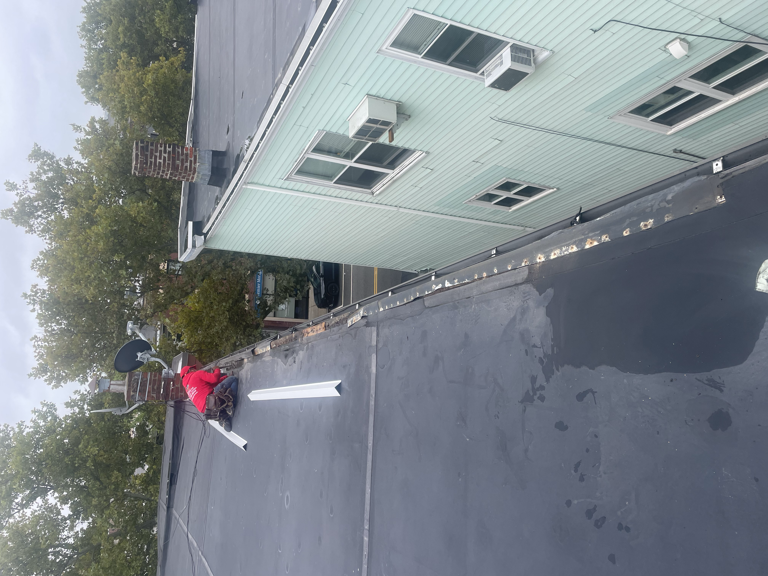 Person in red shirt working on a flat rooftop next to a satellite dish and chimney, with a green building and trees in the background.