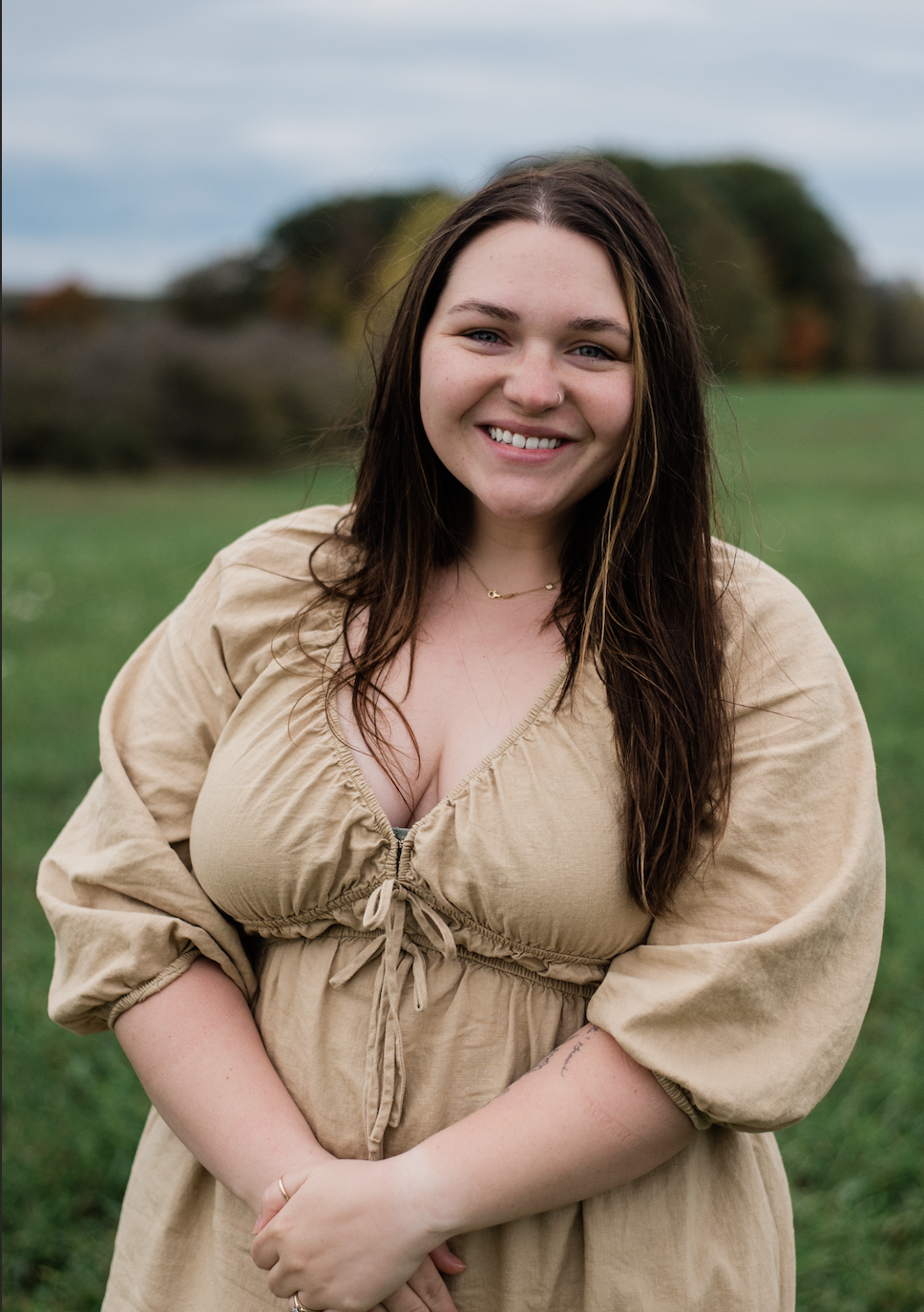 A young woman with long brown hair, smiling, standing outdoors in a grassy field with trees in the background, wearing a beige dress with puffy sleeves.
