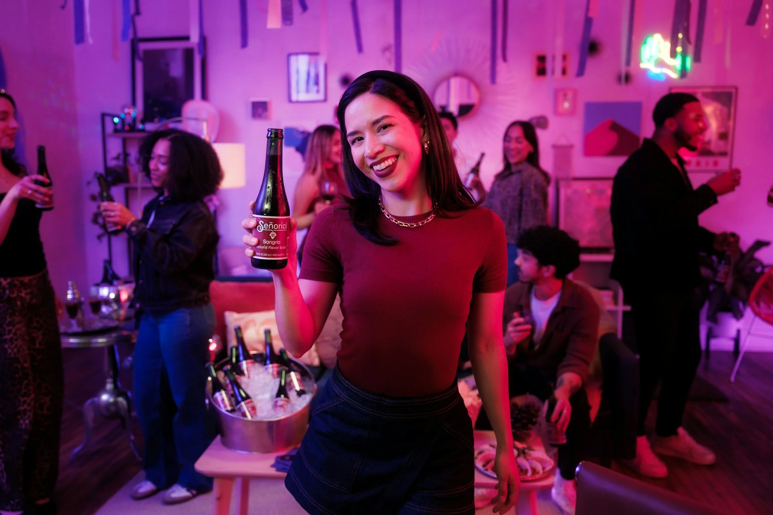 A woman smiling and holding a drink at a party with several other people socializing in a decorated room with colorful lighting.