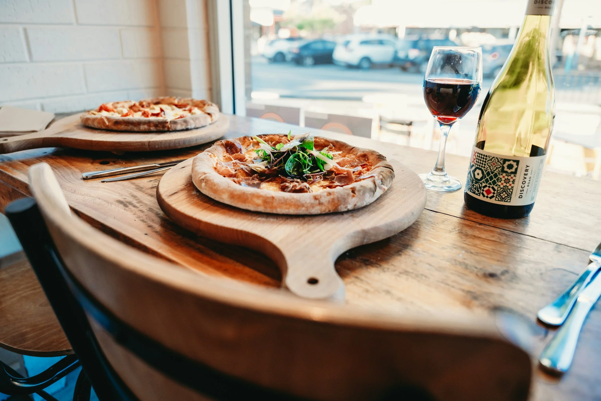 Two pizzas on wooden boards, a glass of red wine, and a bottle of Discovery wine on a rustic wooden table by a window with a parking lot outside.